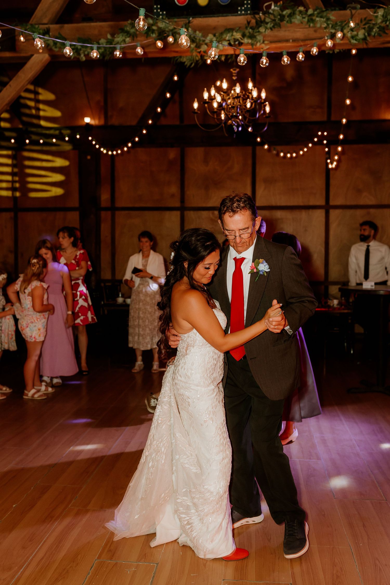 Bride and groom share their first dance in a rustic barn venue adorned with string lights and a chandelier.