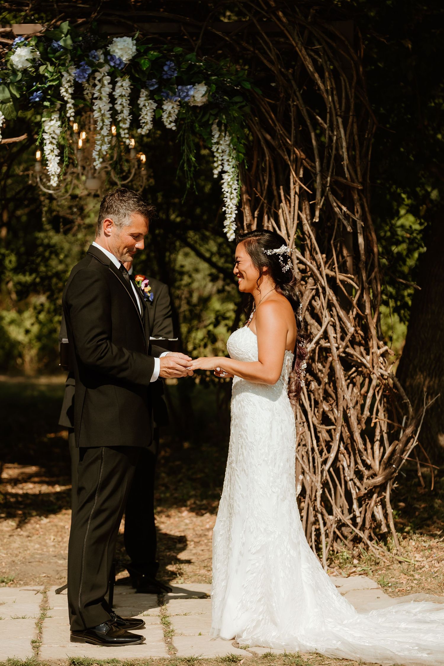 Bride and groom exchange rings outdoors under floral arch with wisteria blooms during sunny wedding ceremony.
