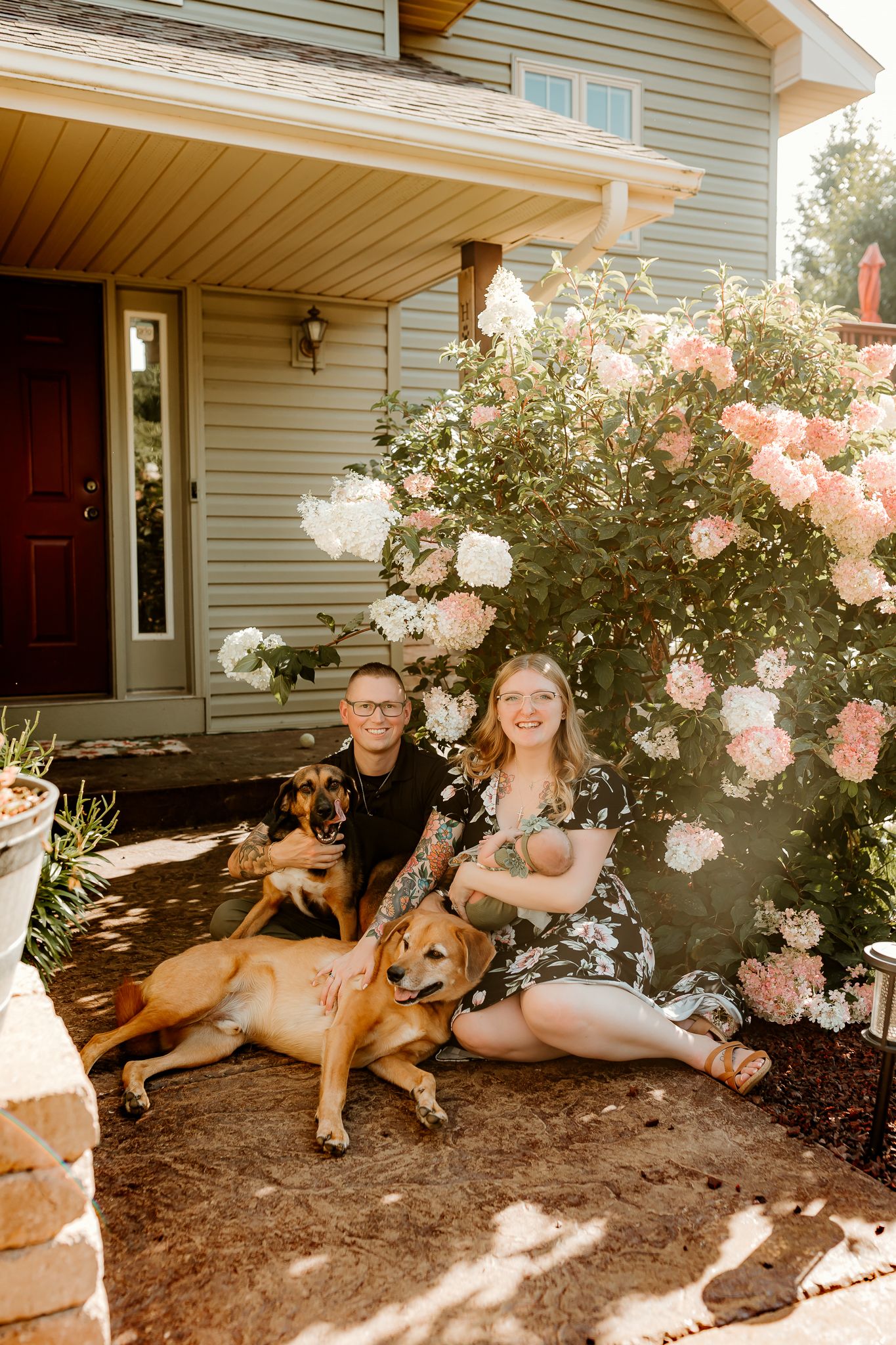 Couple with newborn baby and two dogs sitting in front of a house with blooming white and pink flowers.
