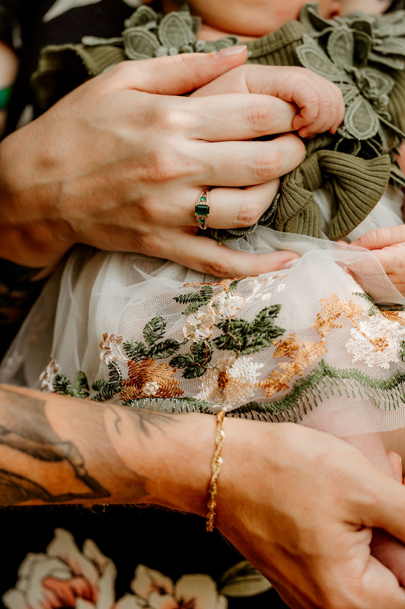 Close-up of hands holding a baby in embroidered dress, woman wearing green gemstone ring, man with floral tattoo and gold bracelet.