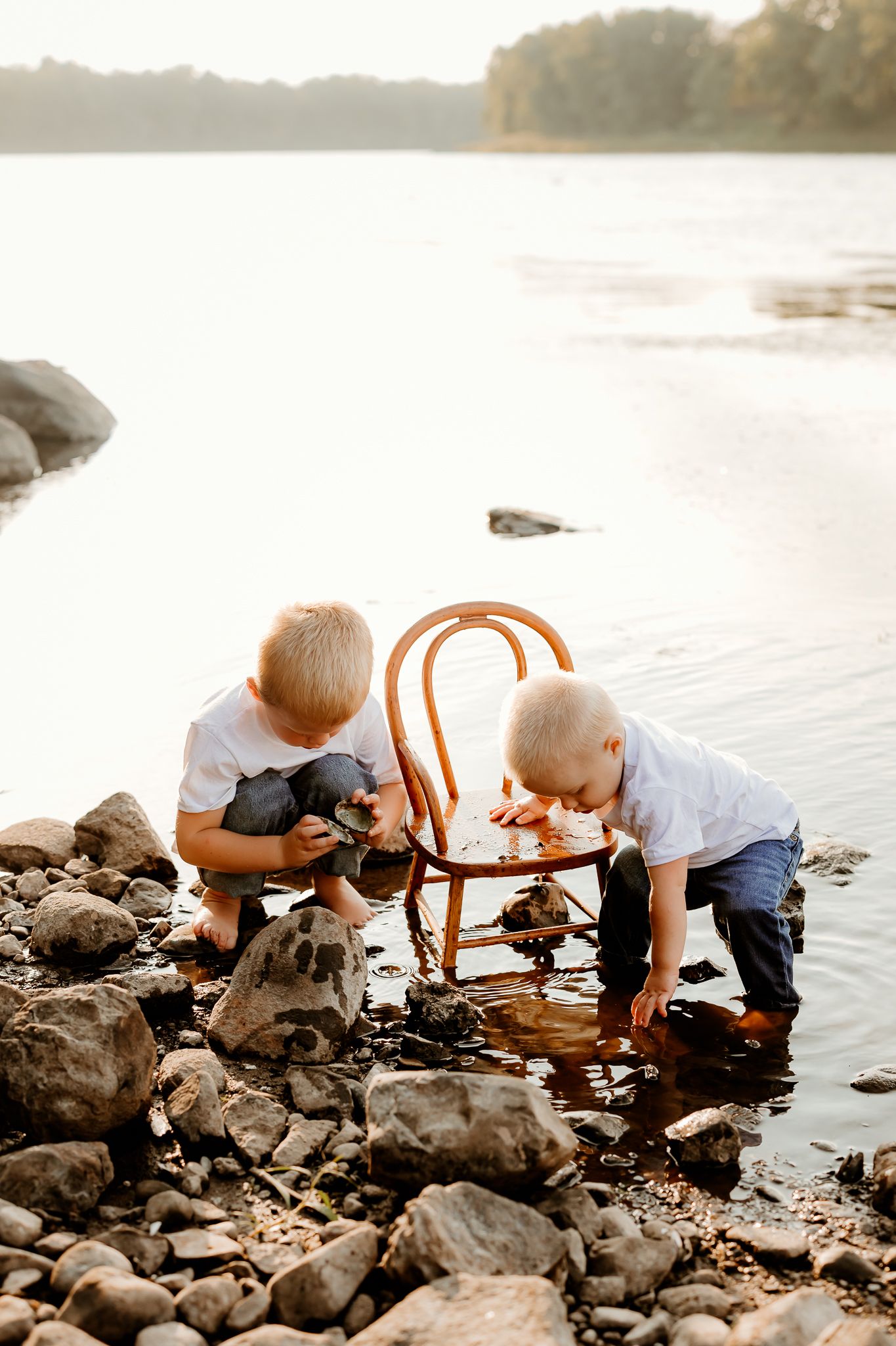 Two young boys in white shirts play with rocks and a wooden chair at the edge of a rocky riverbank at sunset.