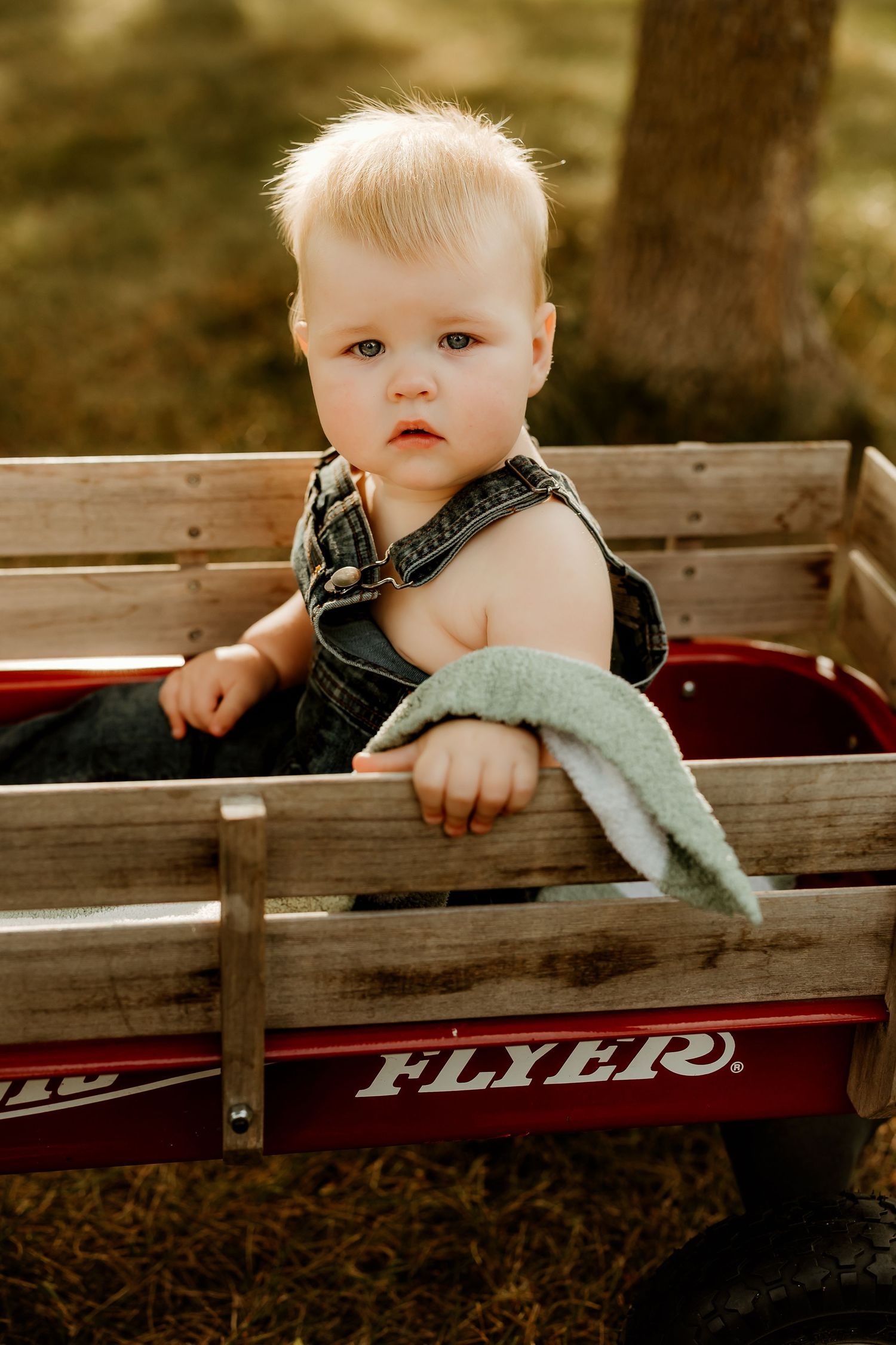 Blonde toddler in denim overalls sitting inside a red Radio Flyer wagon outdoors.