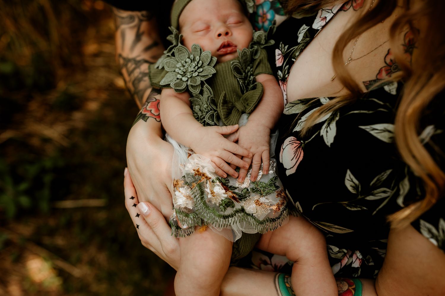 A mother in a floral dress cradles her sleeping newborn baby dressed in a green outfit outdoors.