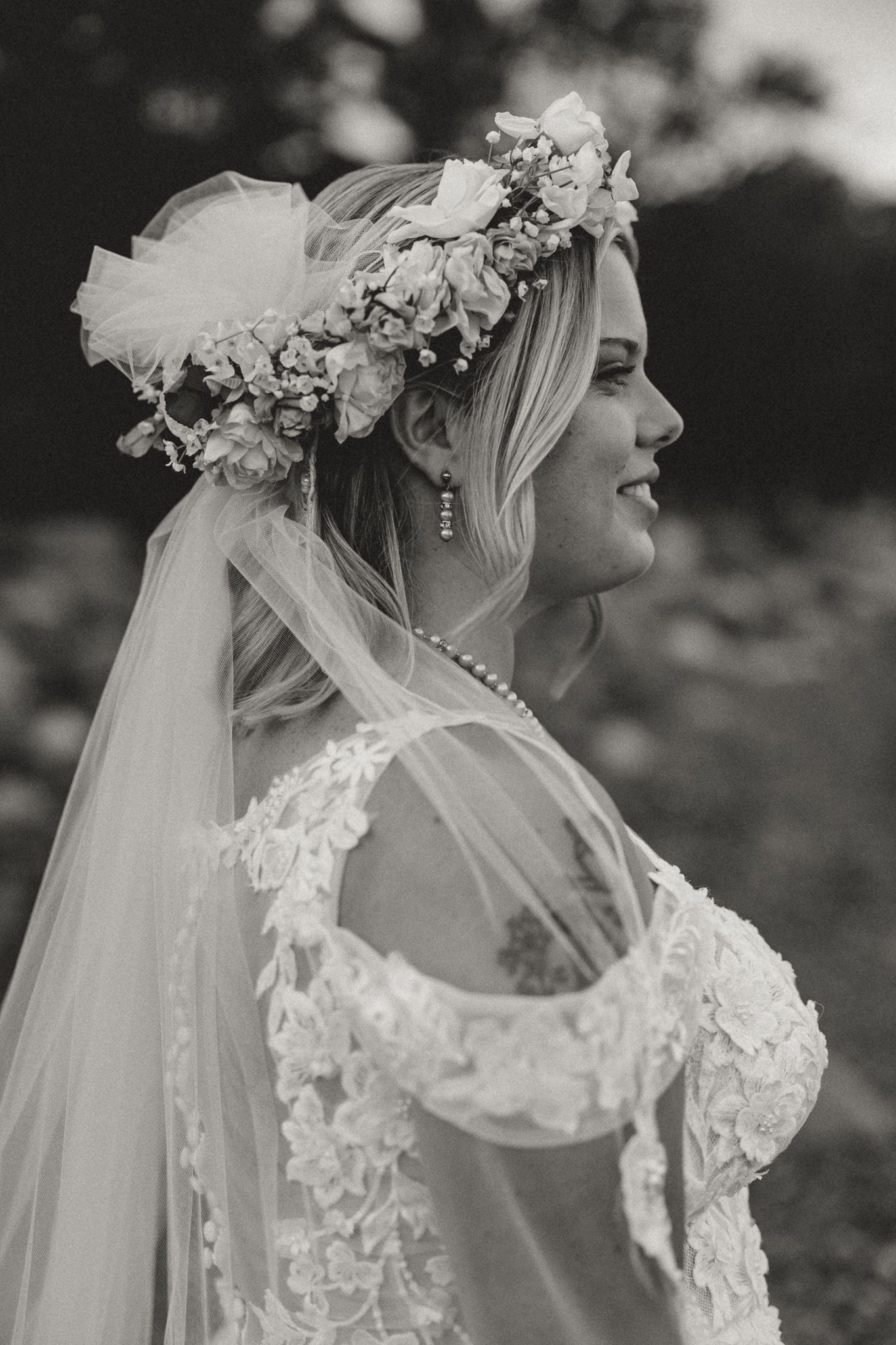 Black and white bridal portrait featuring a smiling bride in lace gown, floral crown, veil, and pearl necklace.