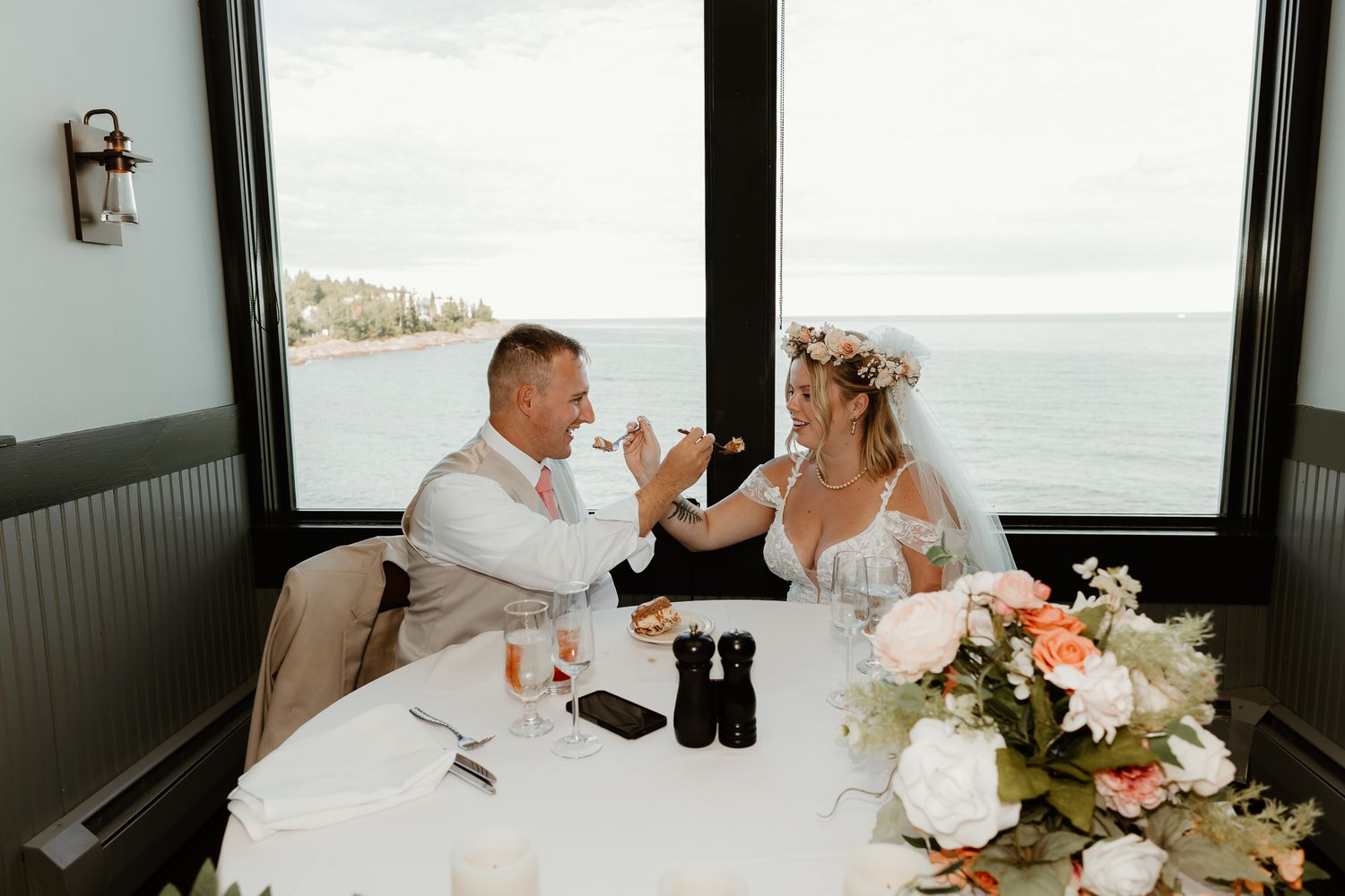 Bride and groom share a toast at a seaside restaurant table adorned with floral arrangements and an ocean view backdrop.