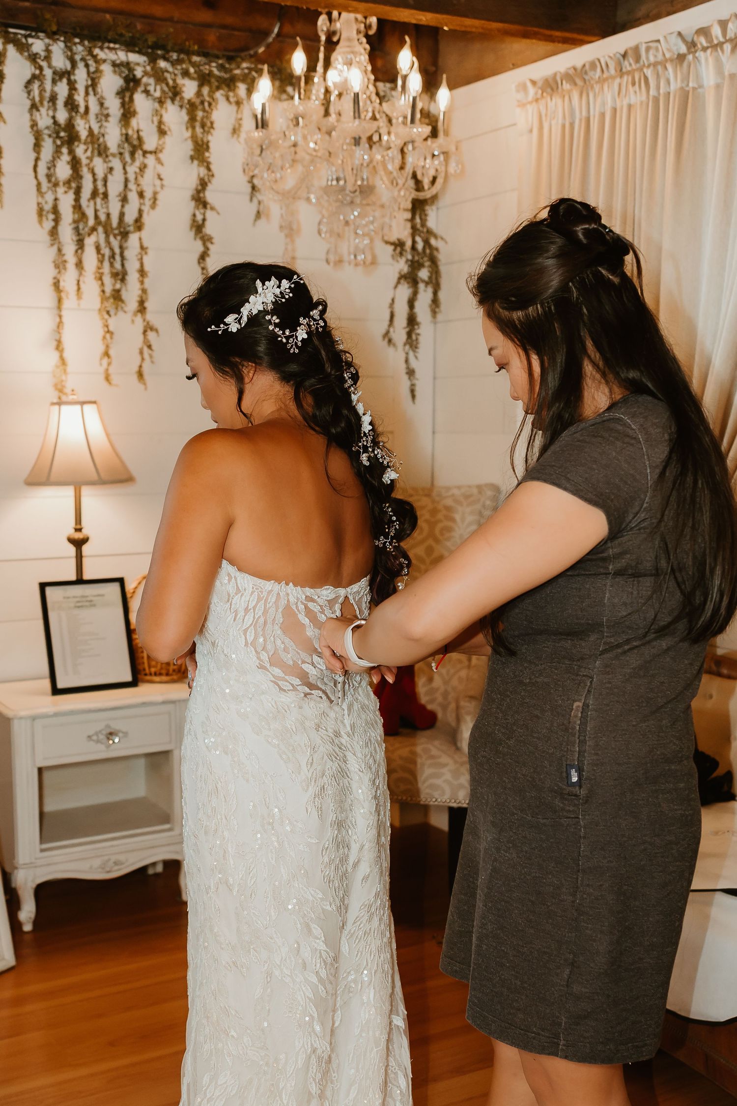 A bride in a beaded gown gets her dress fastened by a helper in a rustic bridal suite with chandelier decor.