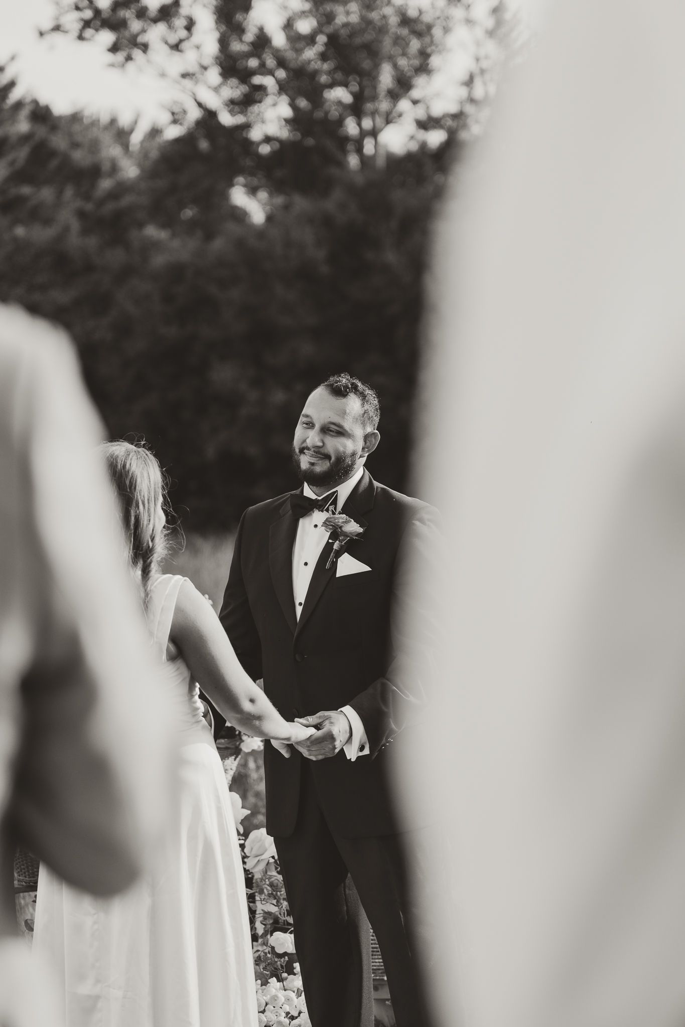 Black and white photo of a smiling groom in a tuxedo holding hands with his bride during an outdoor wedding ceremony.