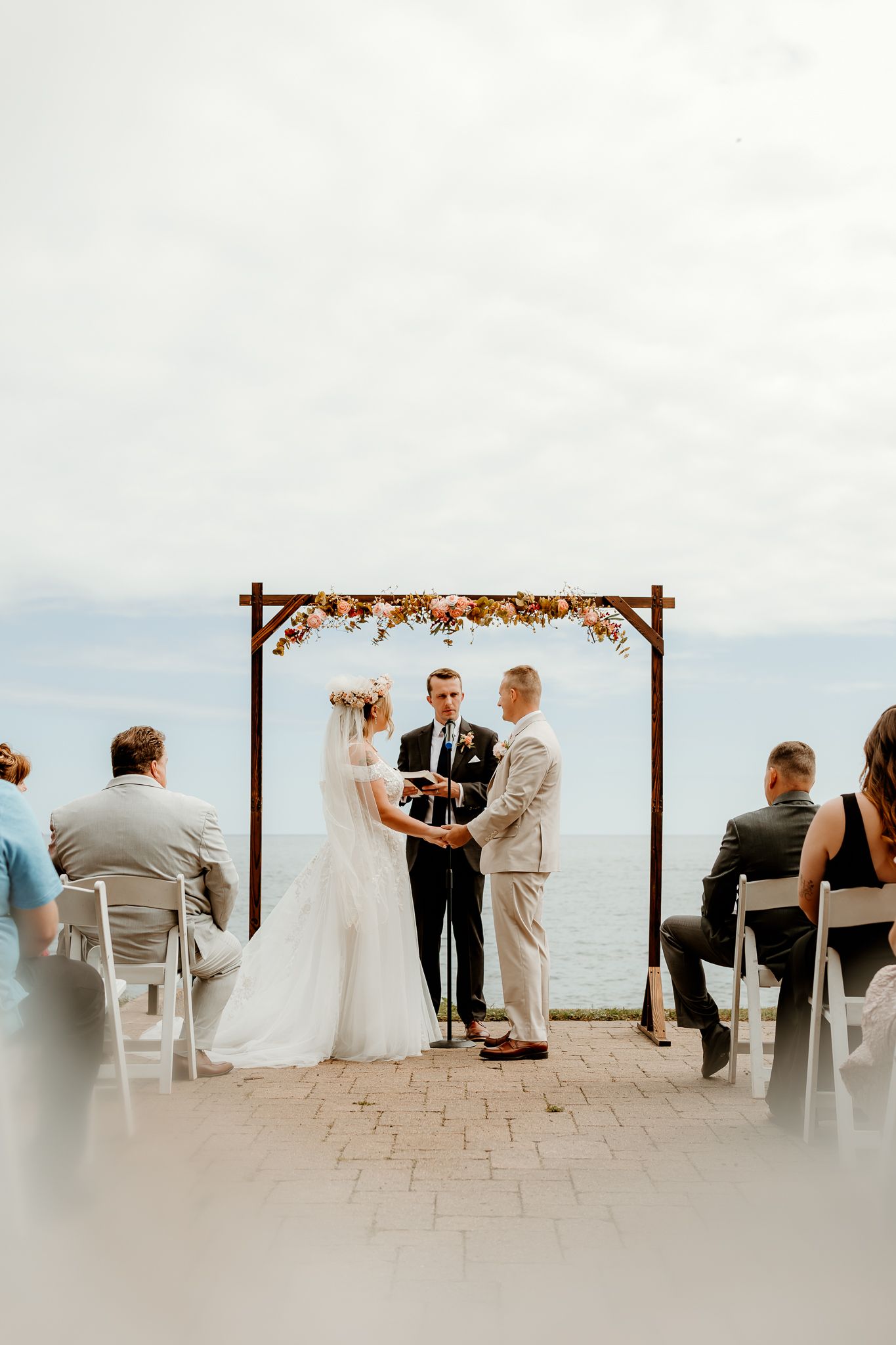 Outdoor wedding ceremony by the ocean with couple exchanging vows under a floral arch, guests seated in white chairs.