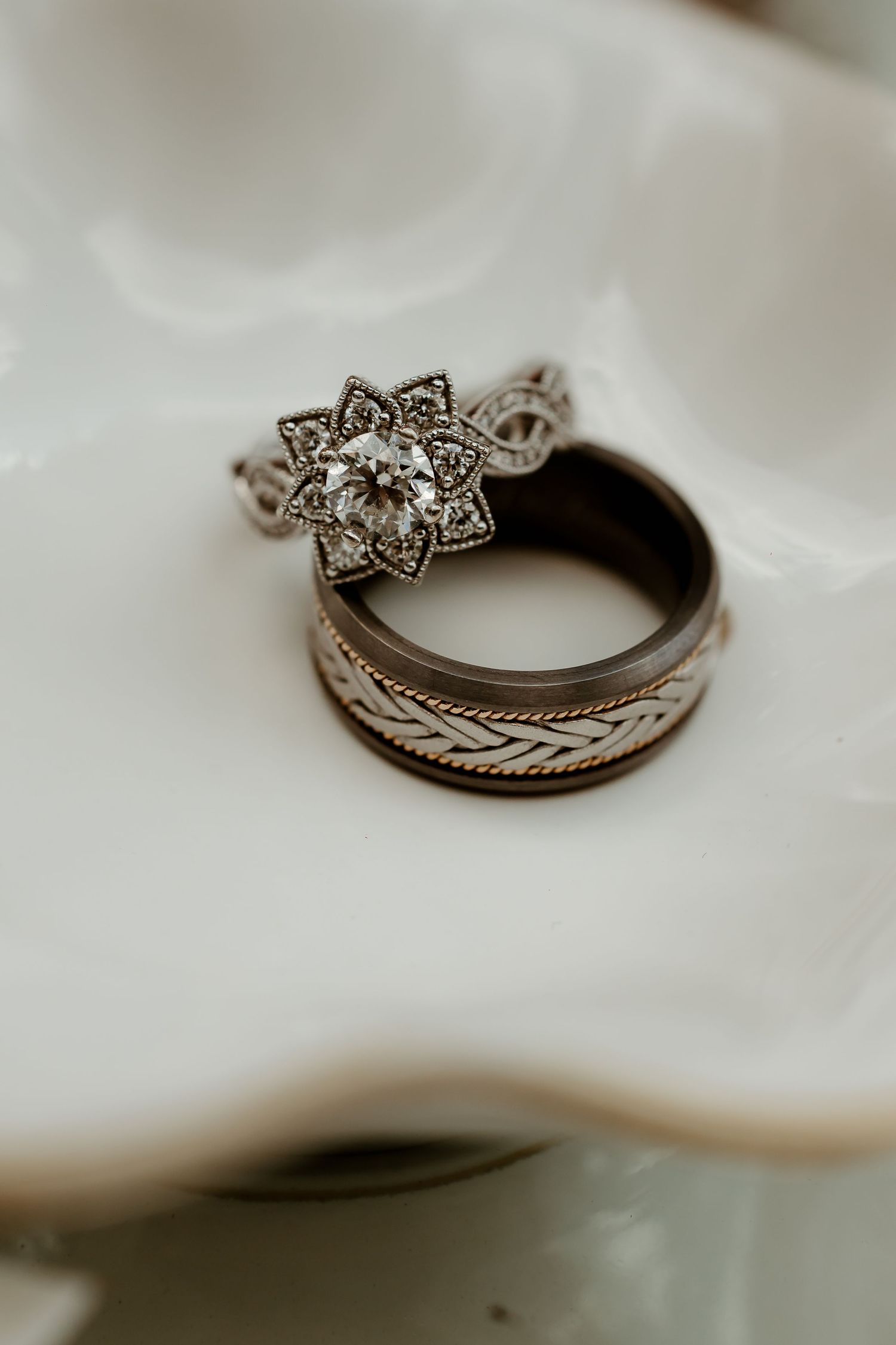 Elegant floral diamond engagement ring paired with engraved wedding band resting on white ceramic dish.