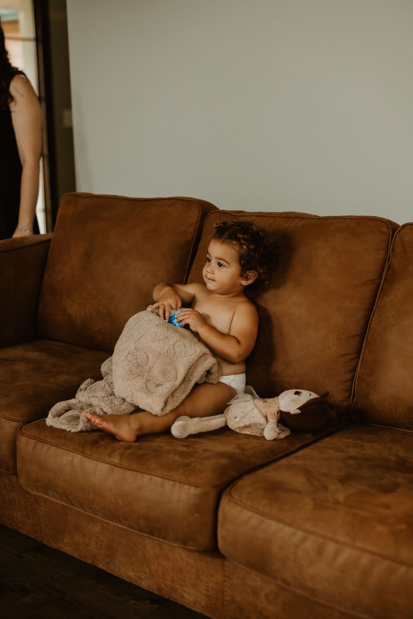 Curly-haired toddler in a diaper sits on a tan leather sofa holding a small toy, with a knit blanket and plush doll beside them.