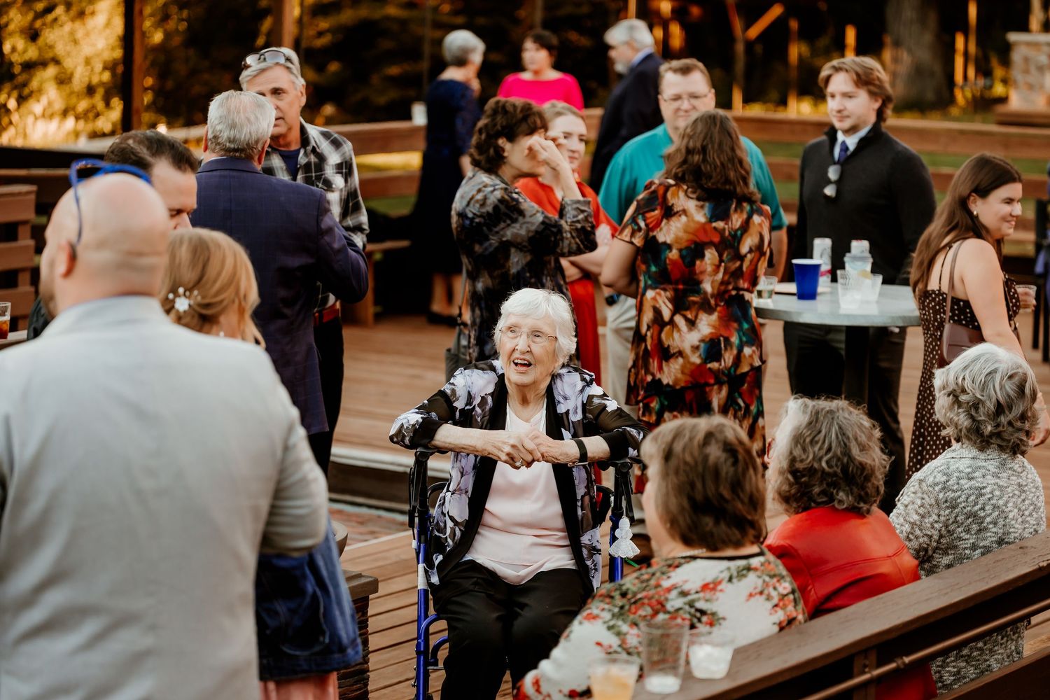 Elderly woman in a wheelchair smiles surrounded by guests mingling at an outdoor wedding reception on a wooden deck.