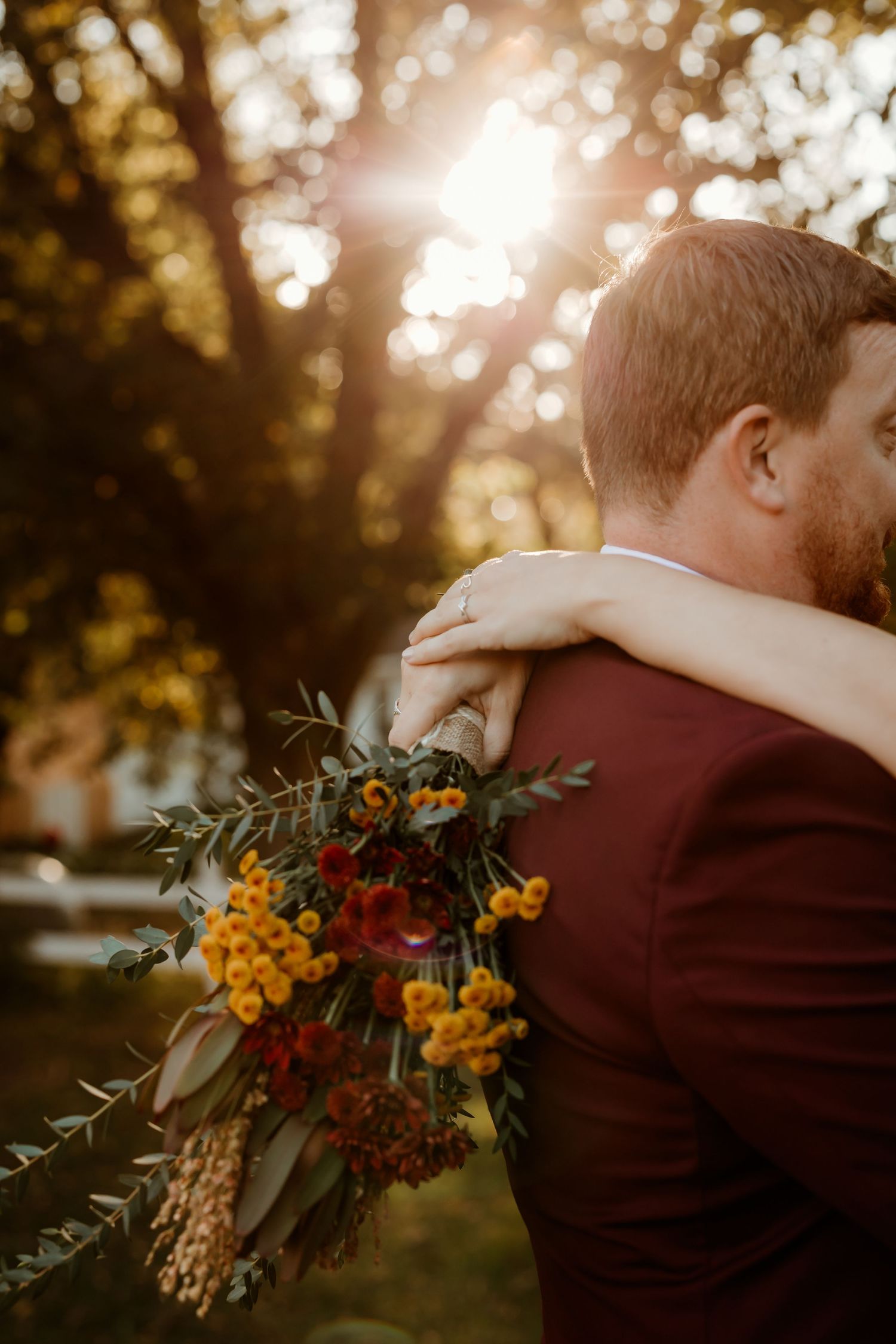 Bride embraces groom in a burgundy suit, holding an autumn bouquet of orange and red flowers with greenery under golden sunset light.