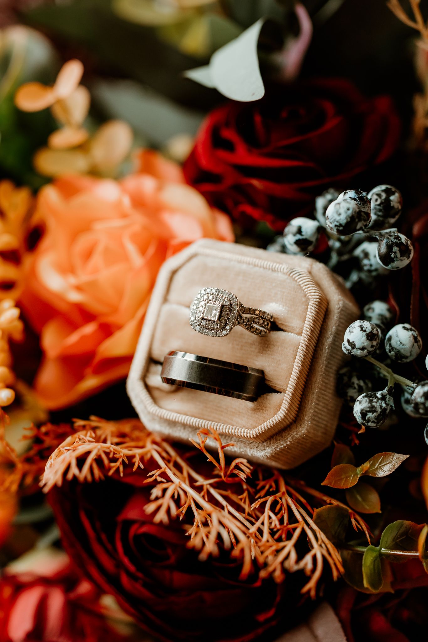 Diamond halo engagement ring and wedding bands in a blush velvet ring box surrounded by orange and red roses and berries.