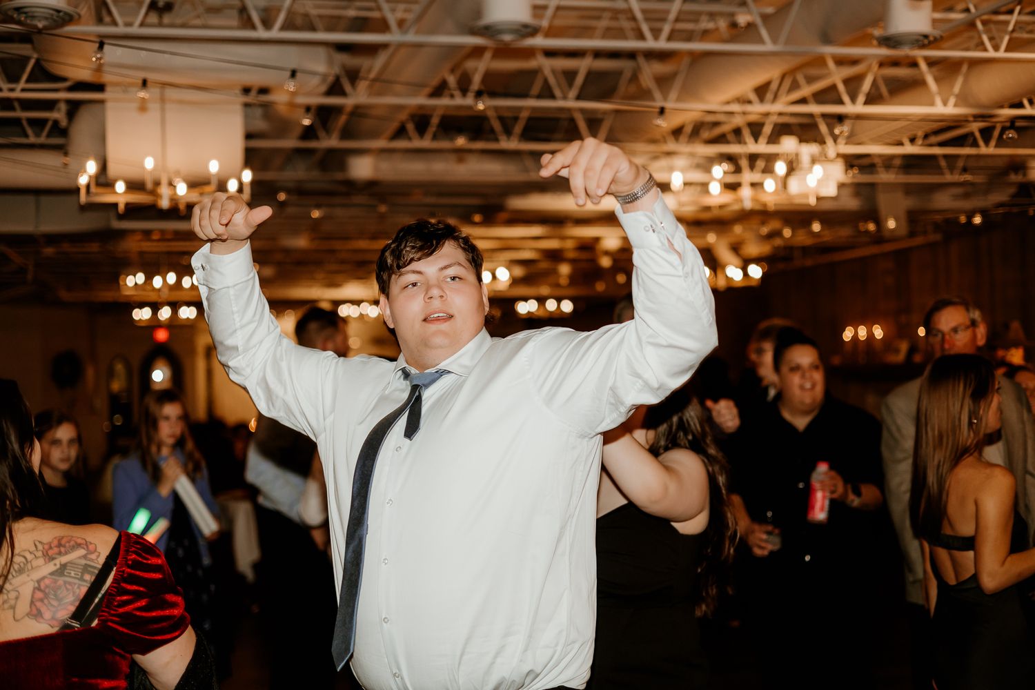 Man in white dress shirt and loosened tie dances with arms raised at a lively indoor party, with guests and string lights in the background.