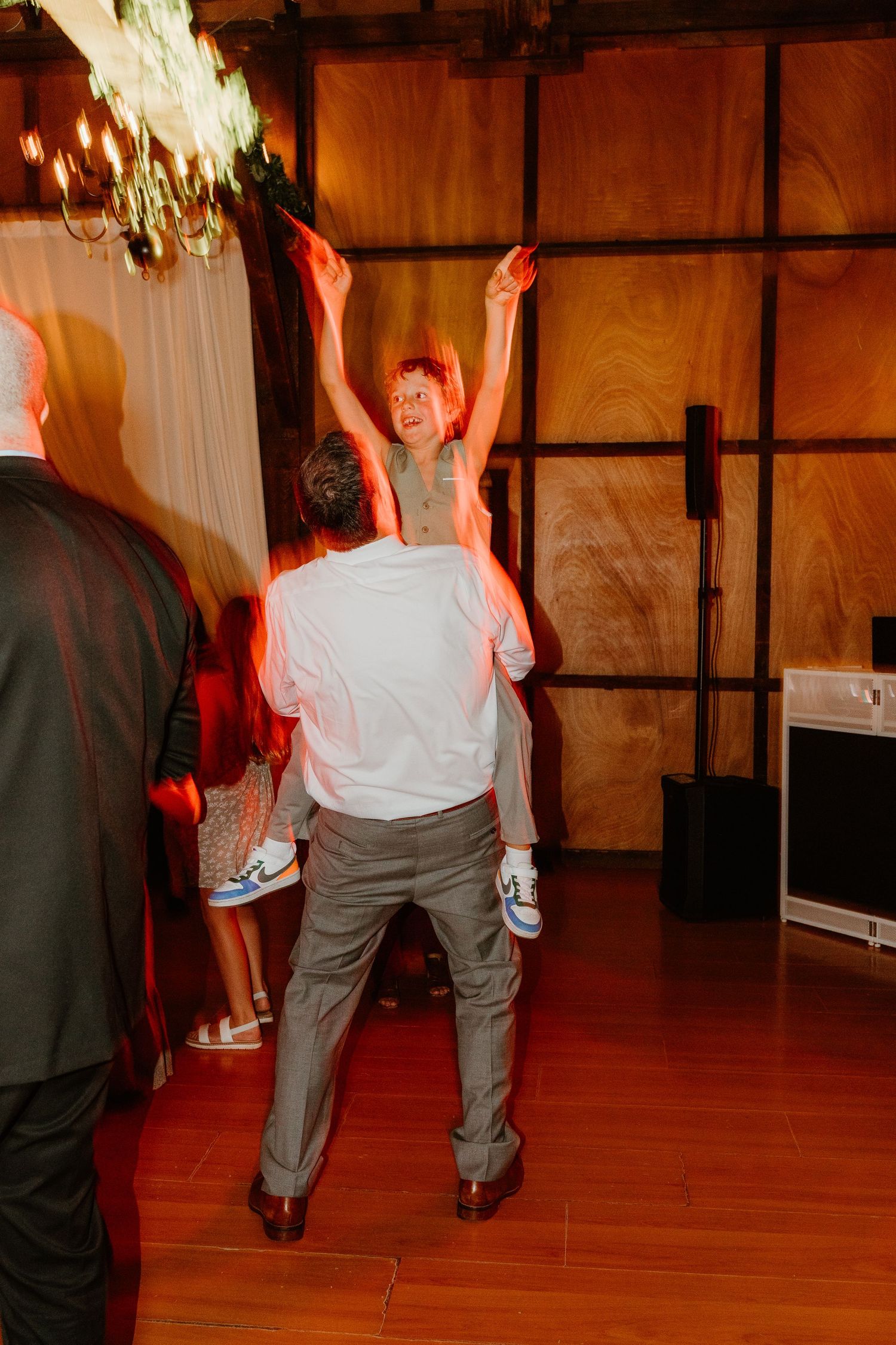 Man in a white shirt lifts a smiling child on his shoulders while dancing at a lively indoor wedding reception with warm lighting.