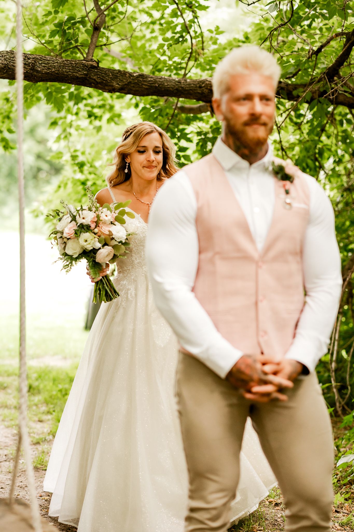 Bride in white gown holding bouquet approaches groom in pink vest during outdoor wedding ceremony under green trees.