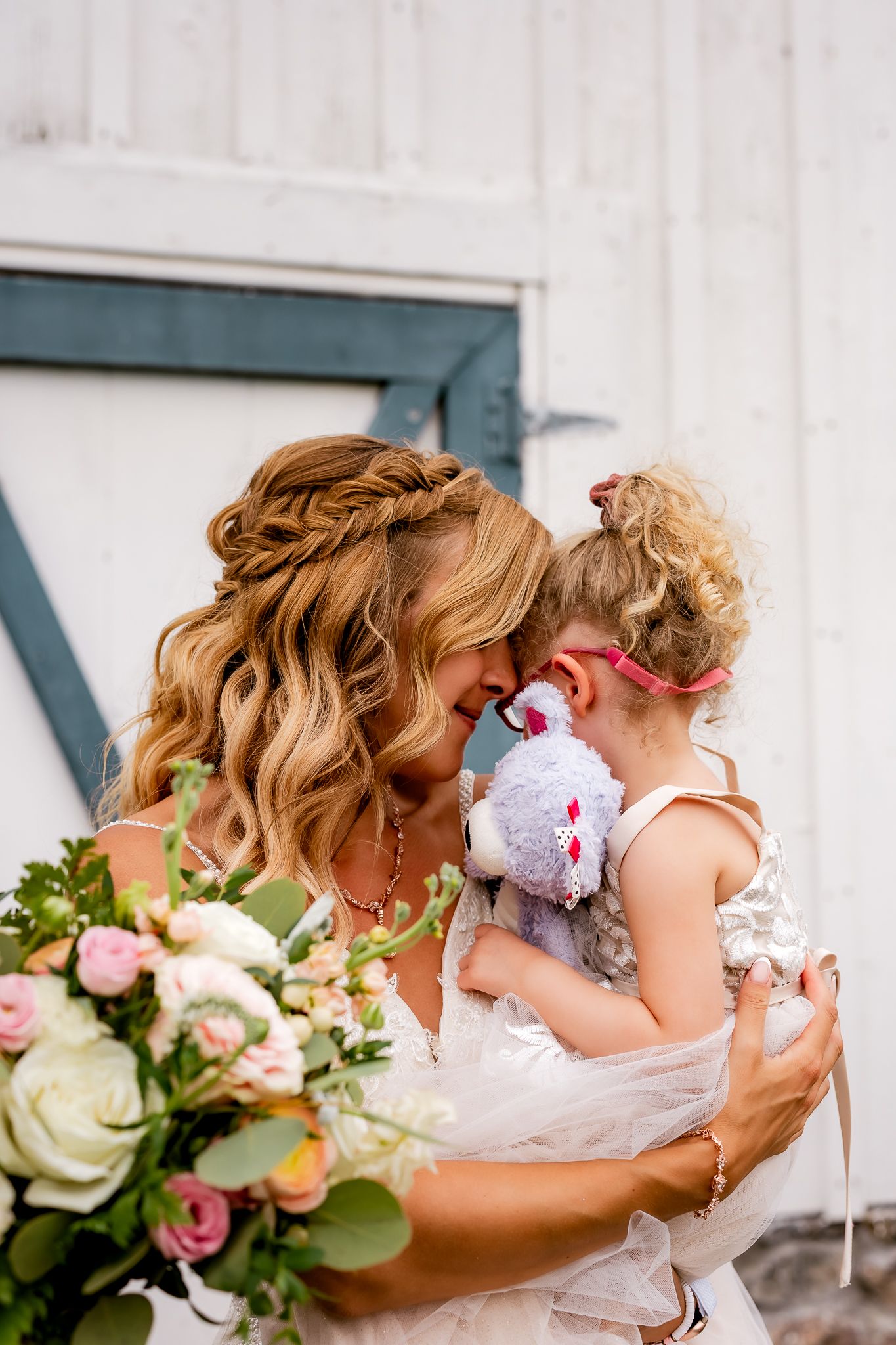 Bride in elegant updo holds young flower girl with stuffed animal, sharing tender forehead touch at barn wedding.