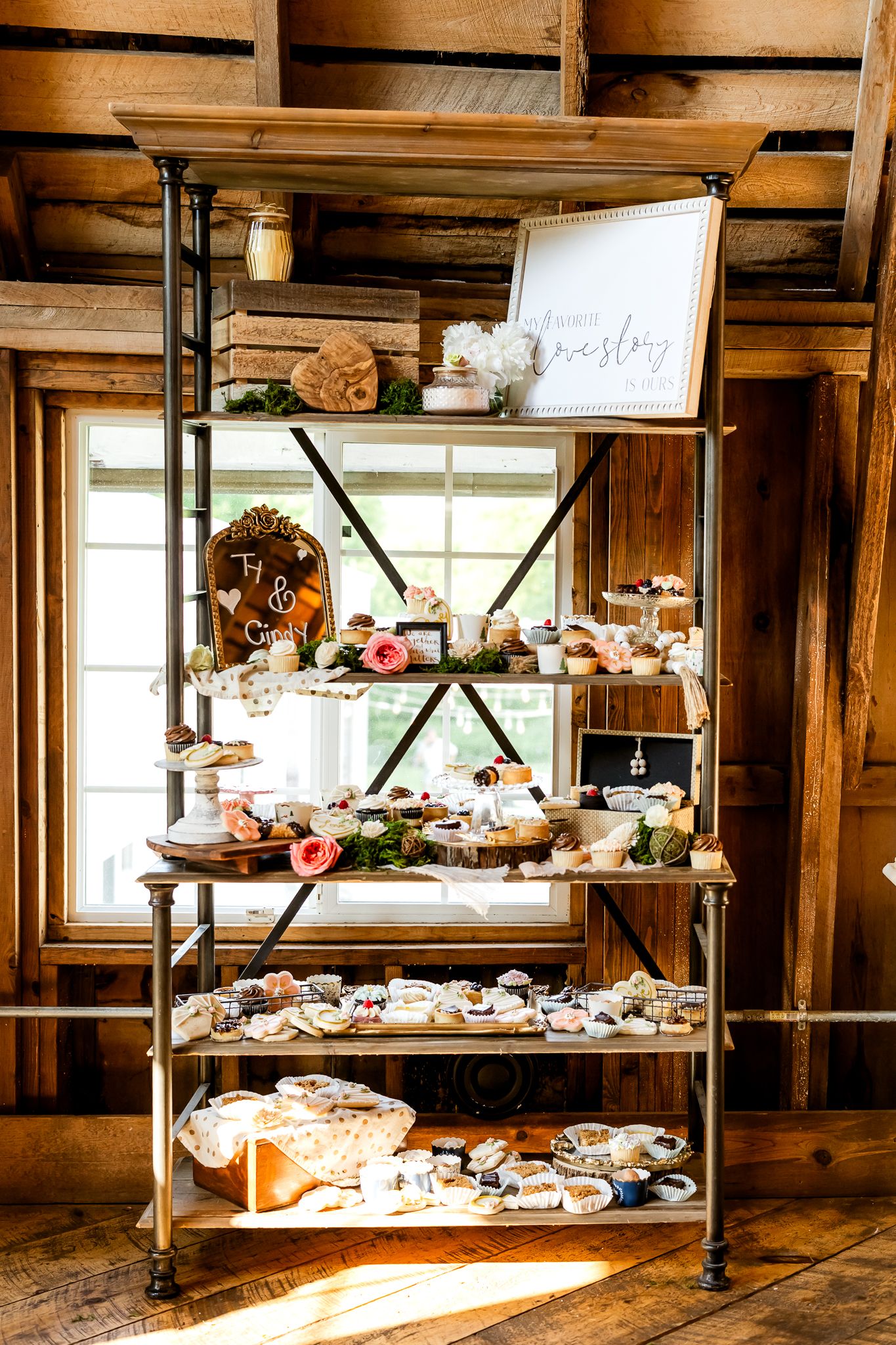 Rustic wedding dessert display shelf with tiered treats, floral accents, and signage in a wooden barn venue.