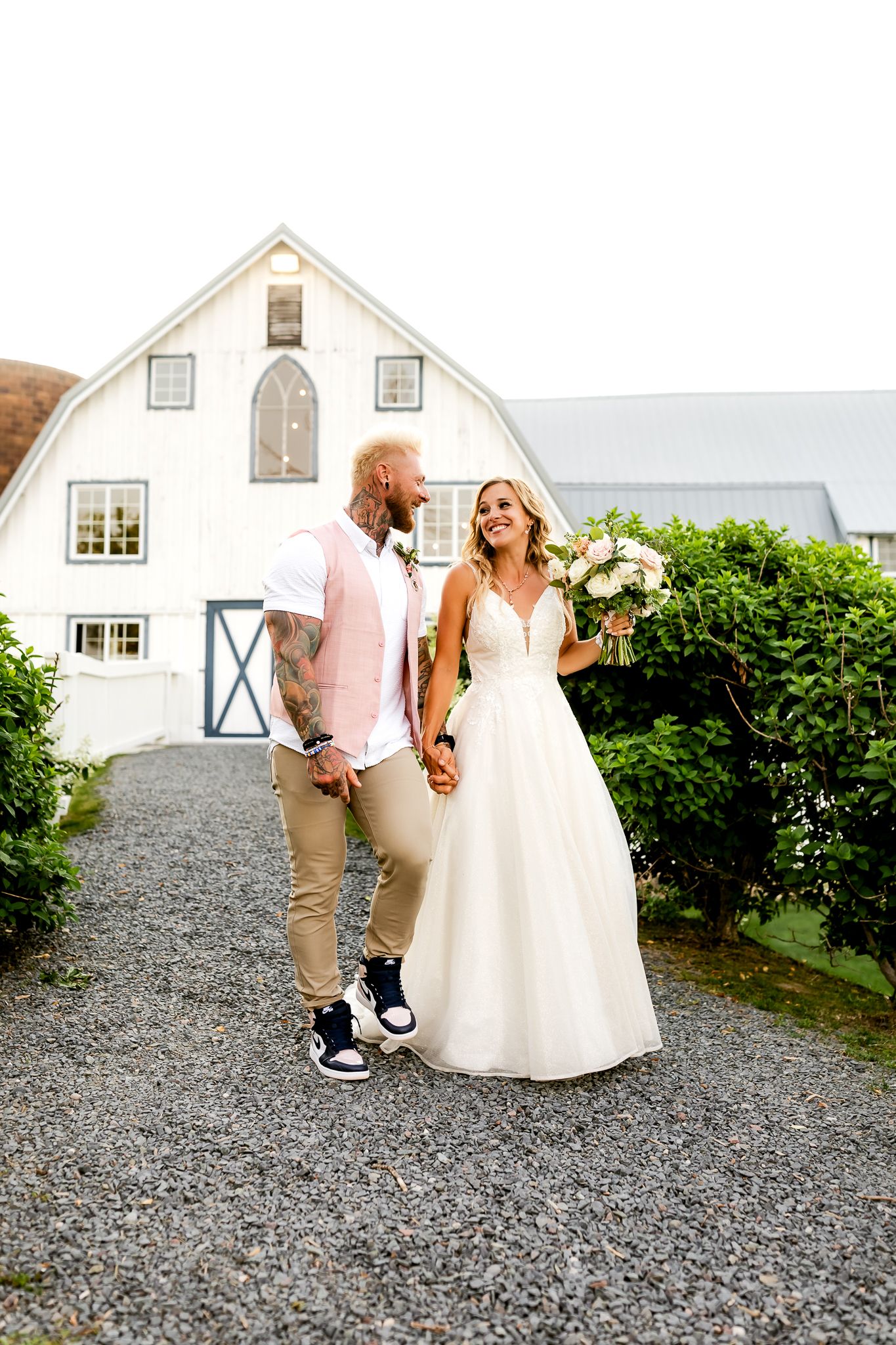 Bride and groom holding hands on gravel path before white barn venue, groom in pink vest, bride in white gown holding bouquet.