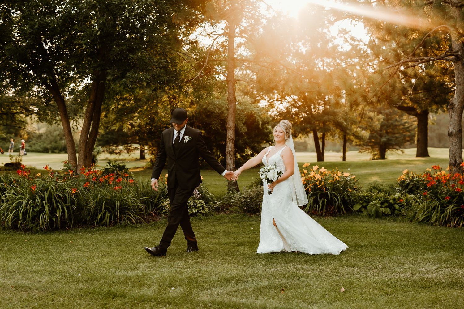 Bride and groom walk hand-in-hand through a sunlit garden with colorful flowers during their outdoor wedding.