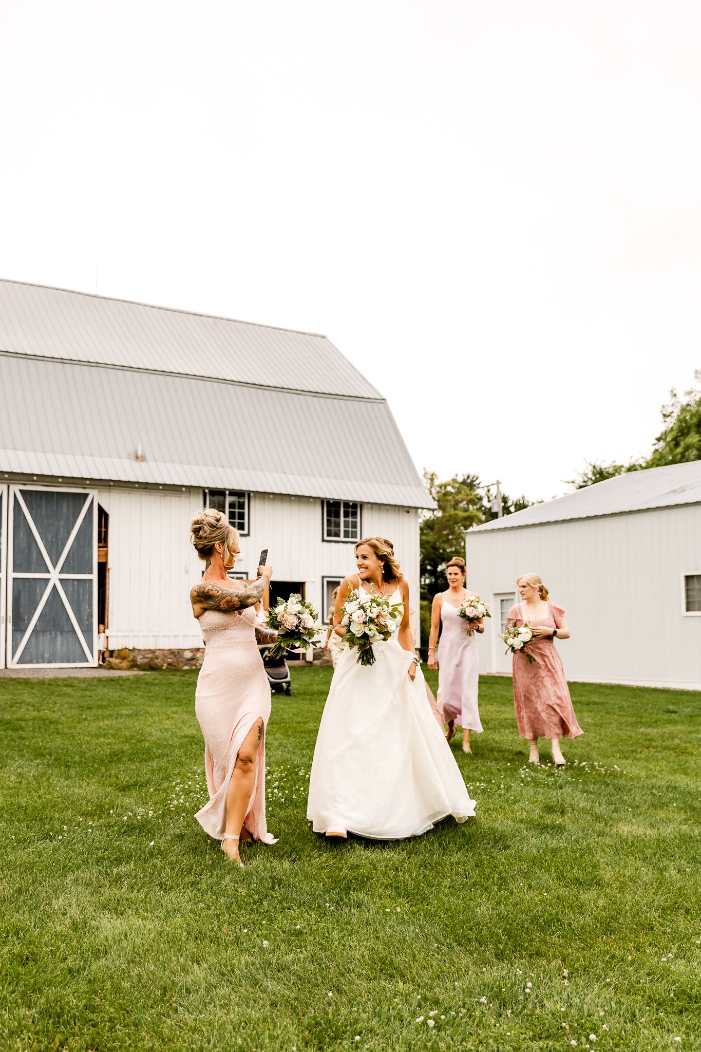Bride in white gown walks with bridesmaids in pink dresses holding bouquets on green lawn near white barn.
