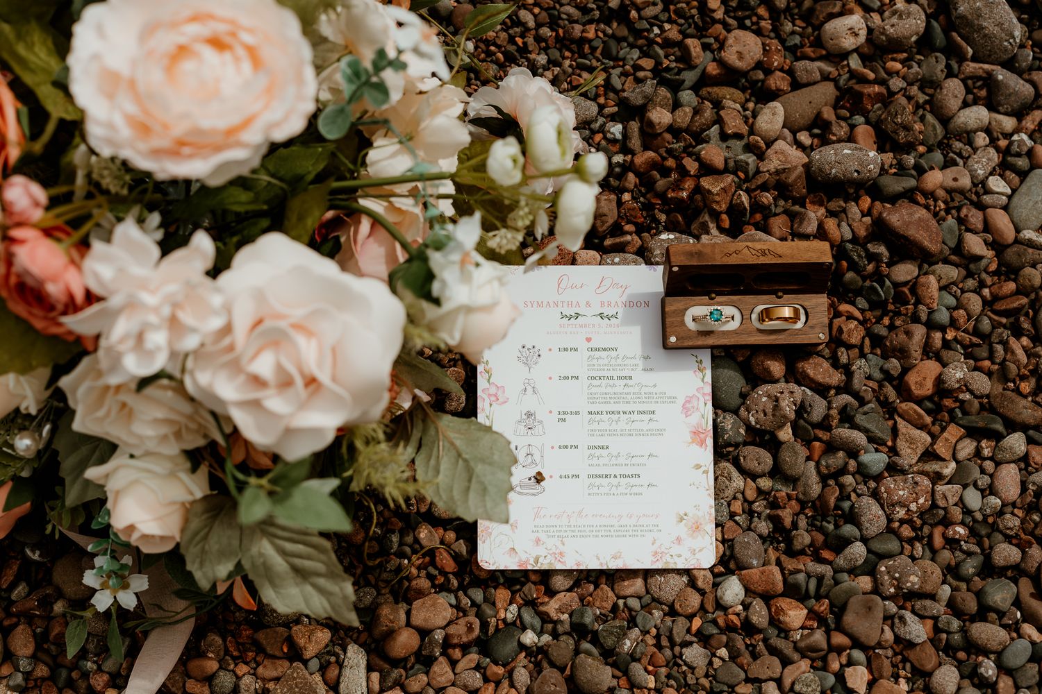 Wedding flat lay with blush roses, ring box with gold bands, and elegant ceremony program on pebble background.