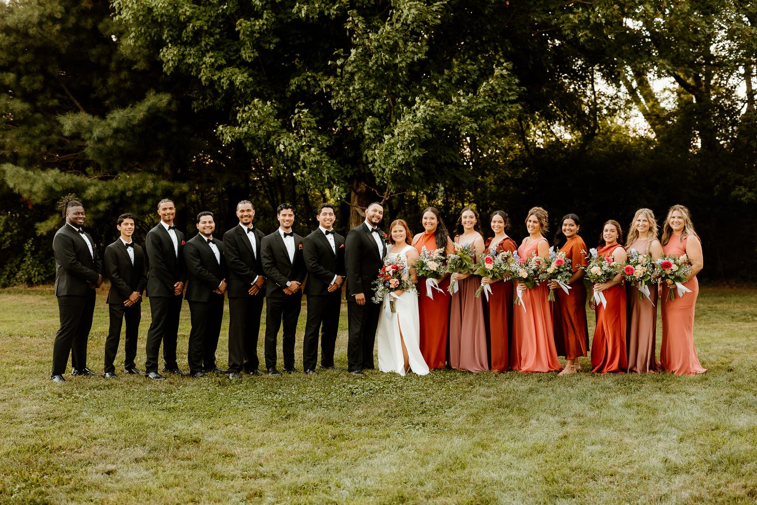 Wedding party posing outdoors, groomsmen in black tuxedos and bridesmaids in red and pink gowns holding bouquets.