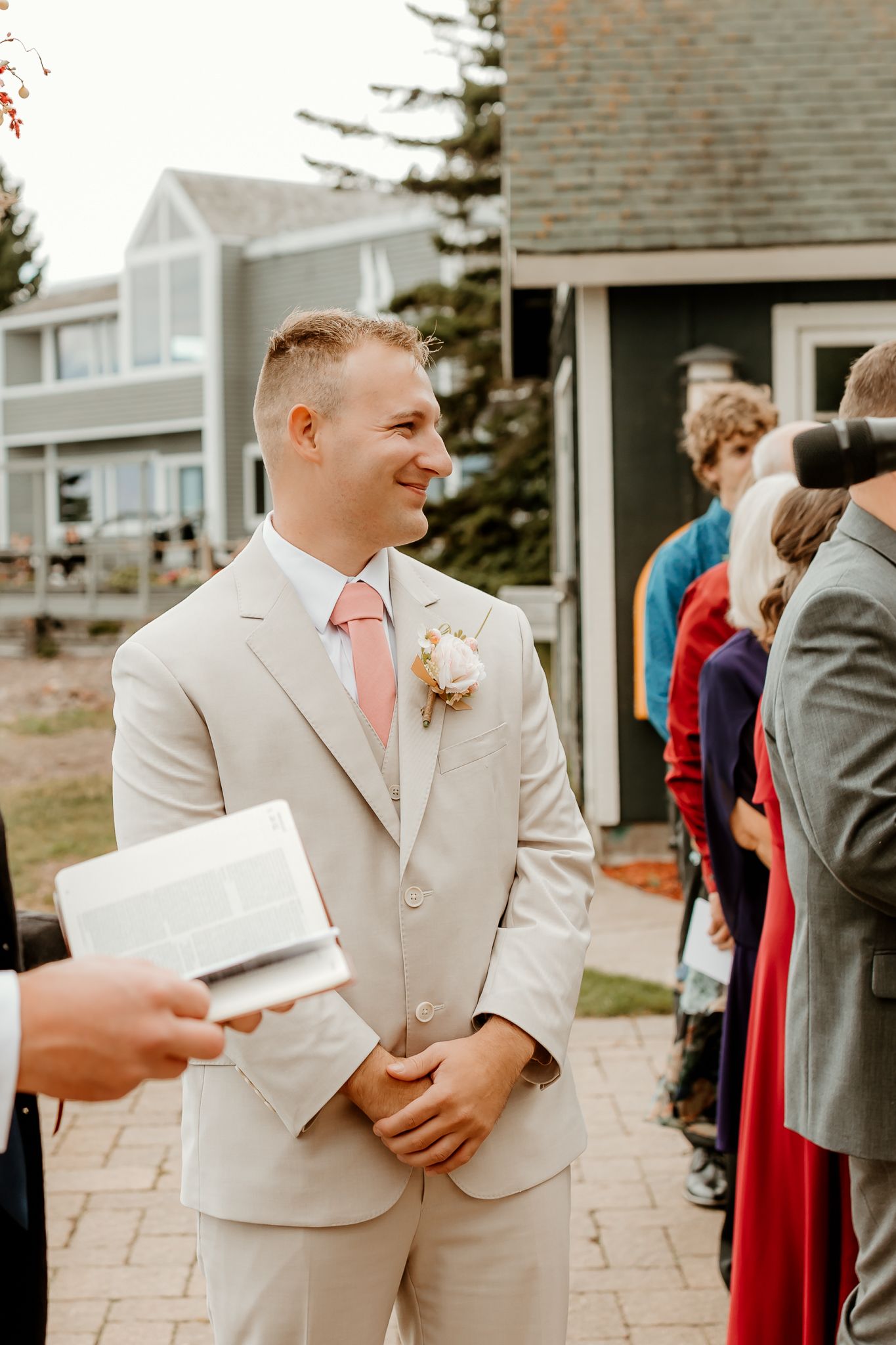 Smiling groom in beige suit with pink tie and boutonniere stands outdoors at wedding ceremony.