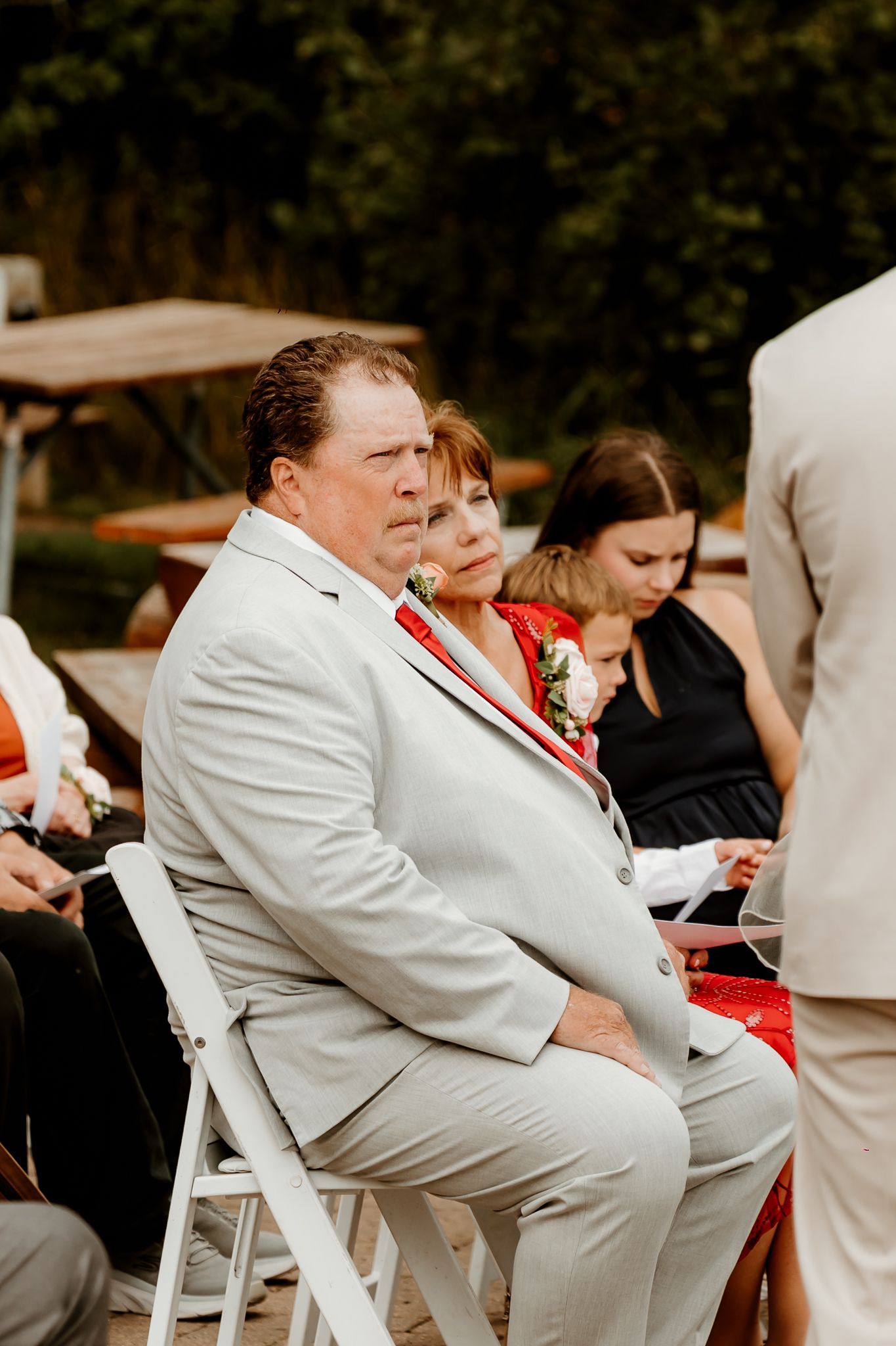 A man in a light gray suit with a red tie sits attentively at an outdoor wedding ceremony alongside other guests.