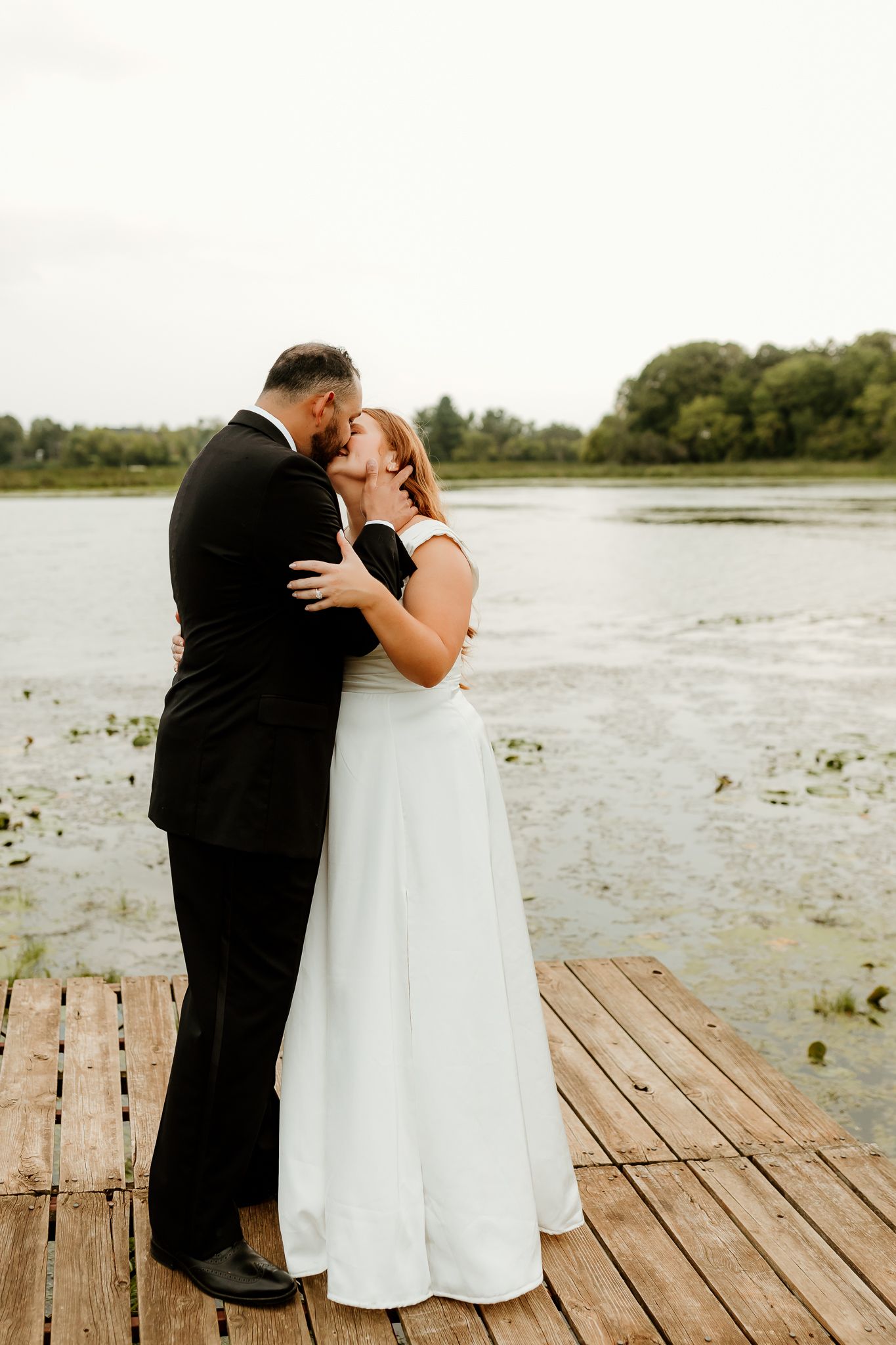 Bride and groom share a romantic kiss on a wooden dock by a serene lake, surrounded by lush greenery.