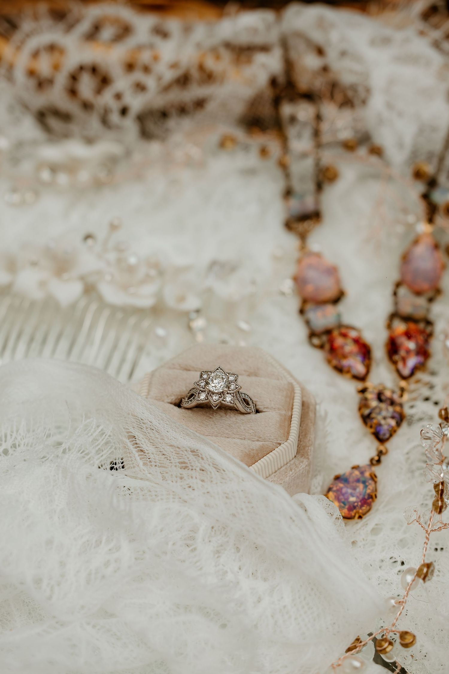 Vintage diamond engagement ring in hexagonal velvet box with lace fabric and colorful gemstone necklace.