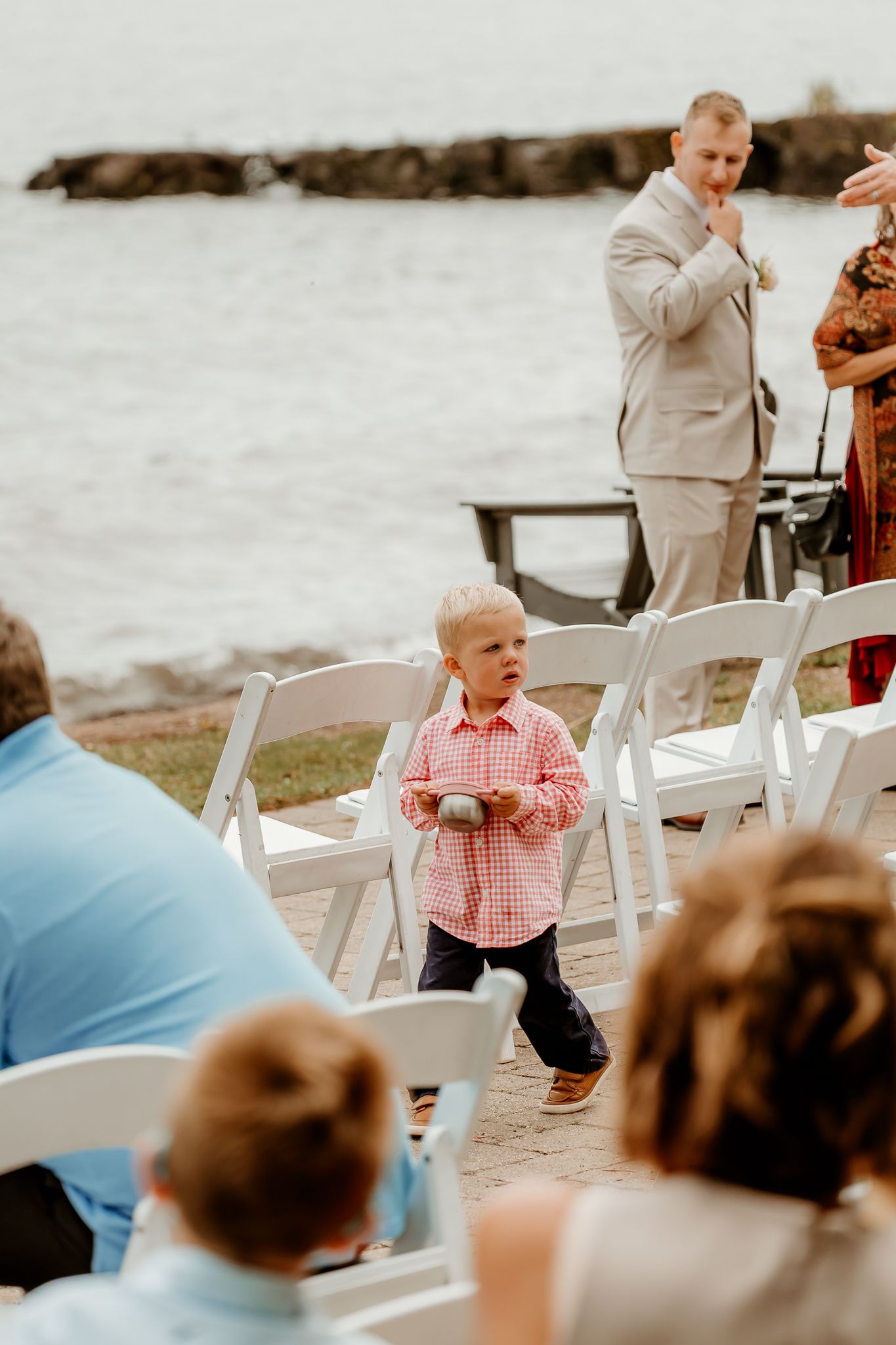 Young ring bearer in pink shirt carries pillow at outdoor waterfront wedding ceremony with white chairs.