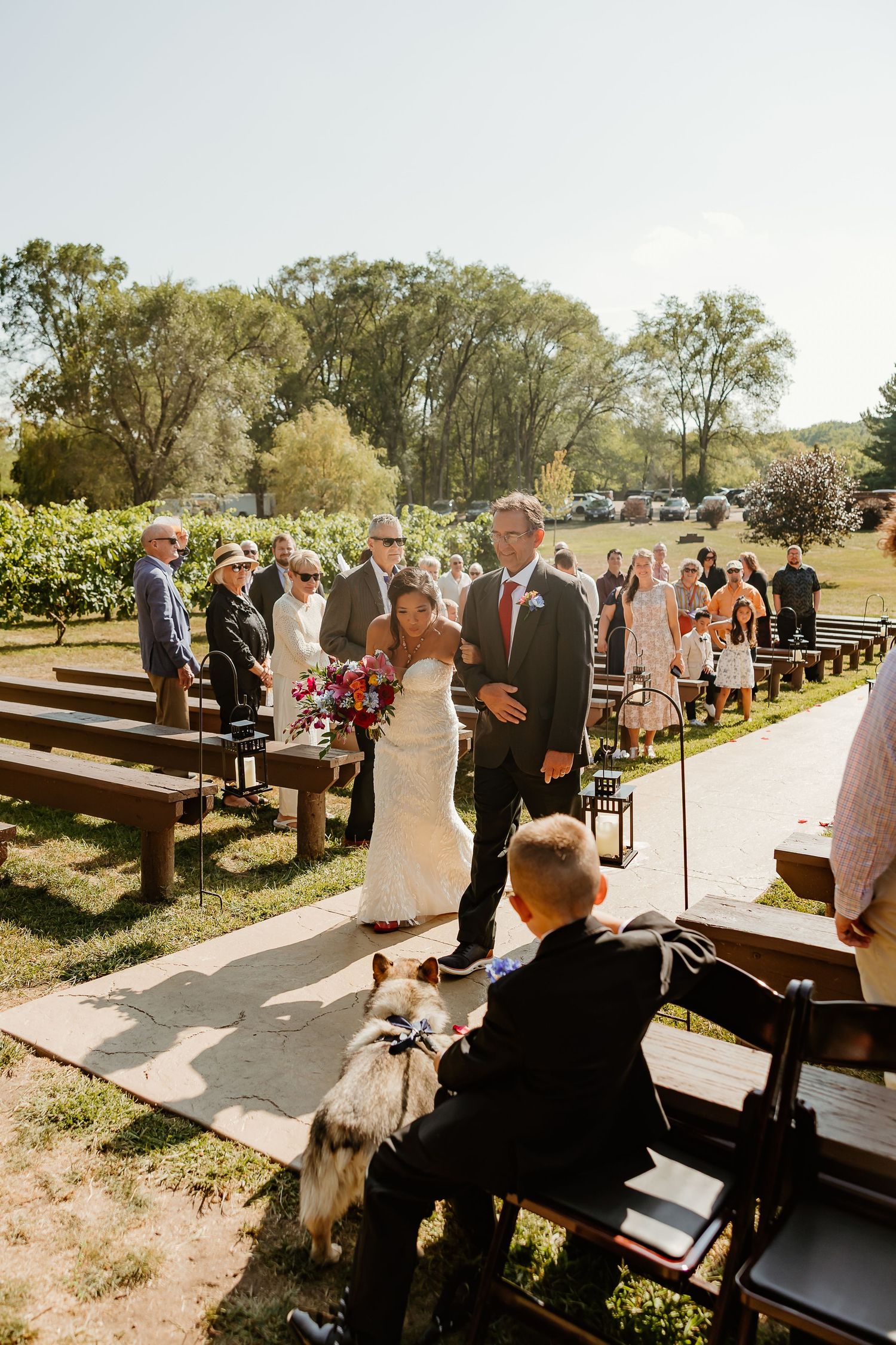 Bride and groom walk down outdoor wedding aisle flanked by wooden benches and guests on a sunny day.