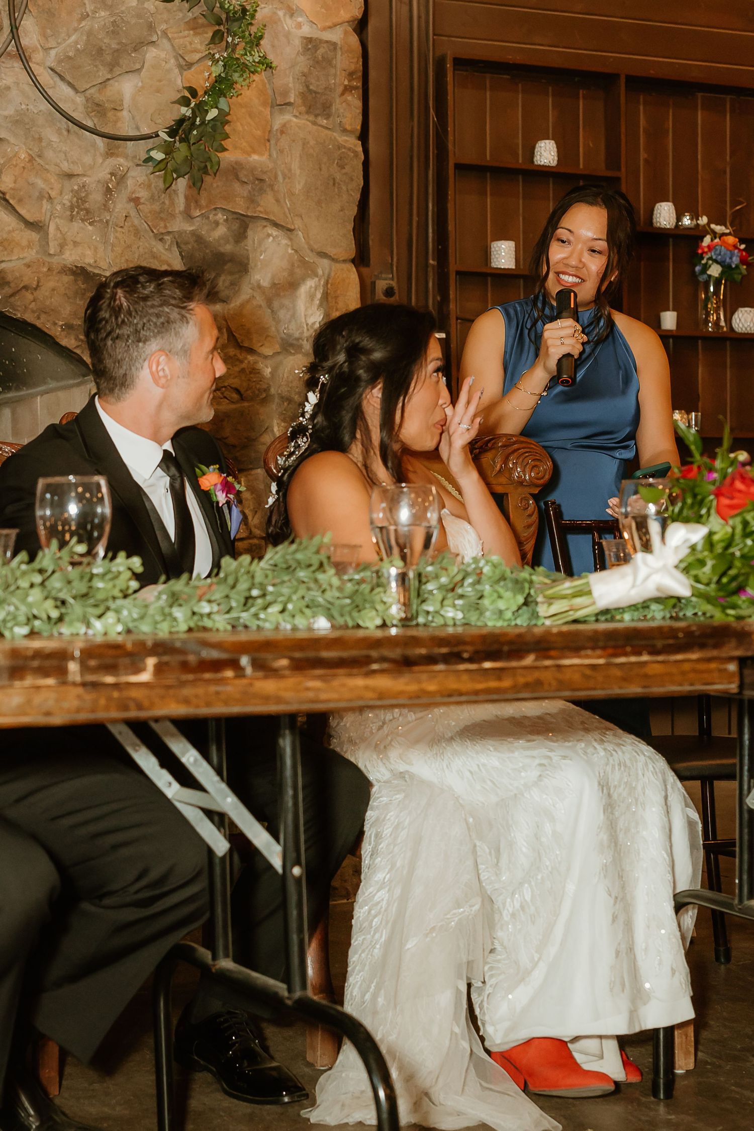 A bridesmaid gives a heartfelt toast with a microphone at a rustic wedding reception while the bride and groom smile.