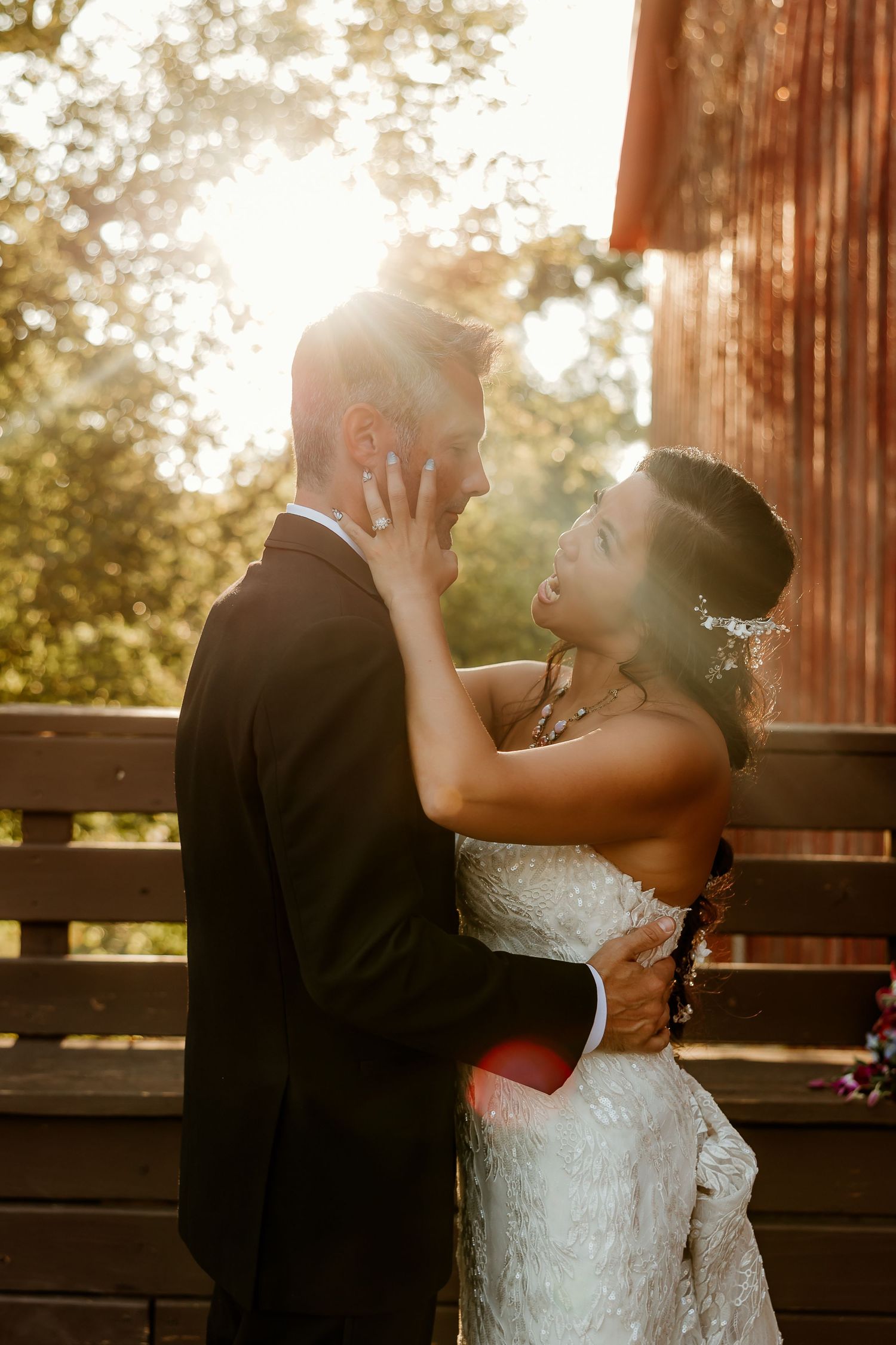 Bride and groom share a romantic embrace at golden hour, with warm sunlight glowing behind them near a rustic barn.