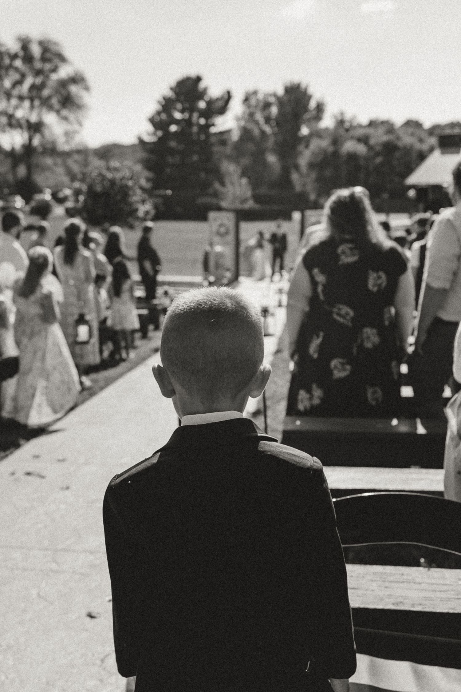 A young boy in a suit sits on a bench watching an outdoor wedding ceremony in a black and white photo.