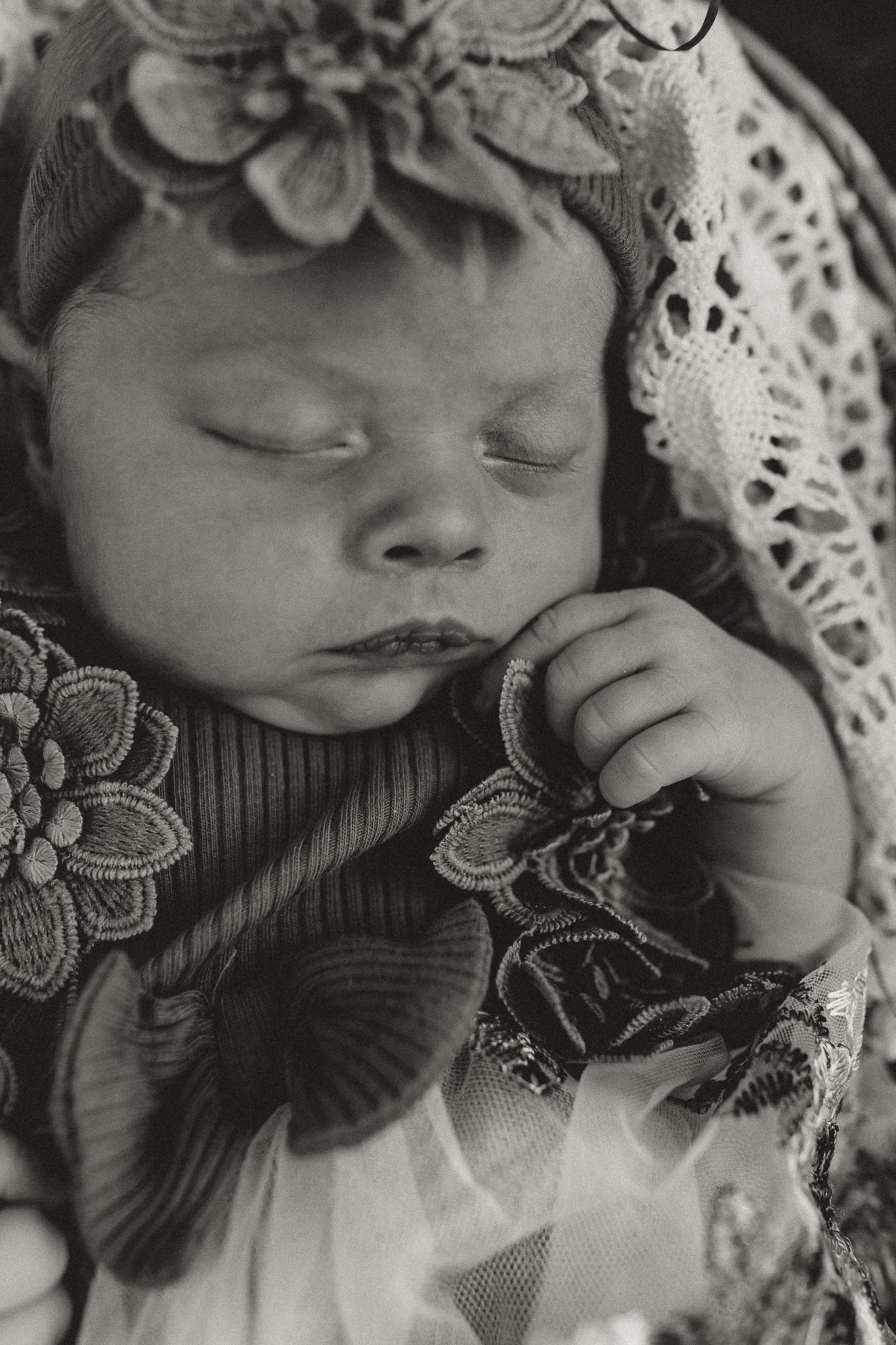 Black and white photo of a sleeping newborn baby girl wearing a floral headband and lace outfit.