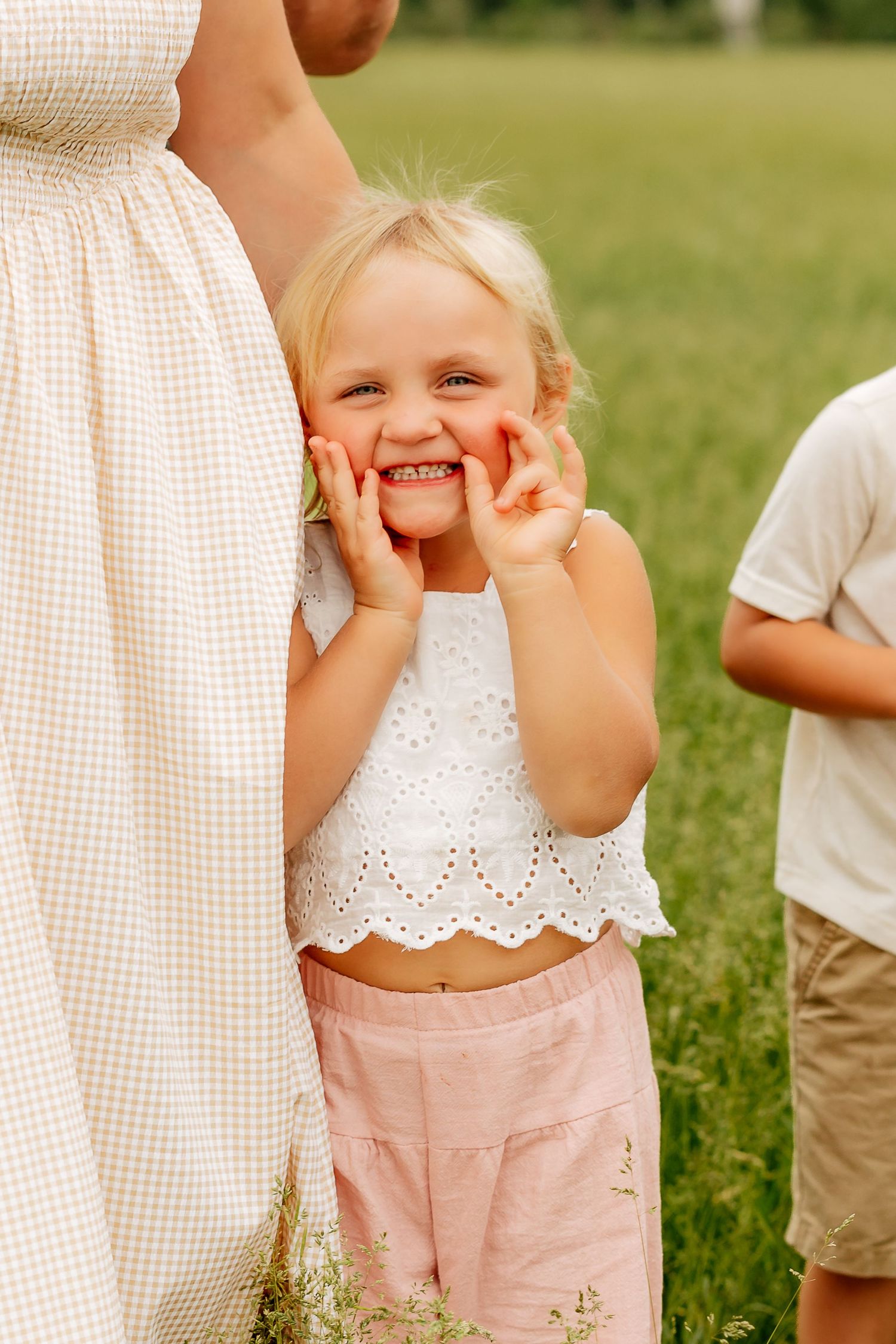 Adorable blonde toddler girl smiling with hands on cheeks, wearing white top and pink shorts outdoors.