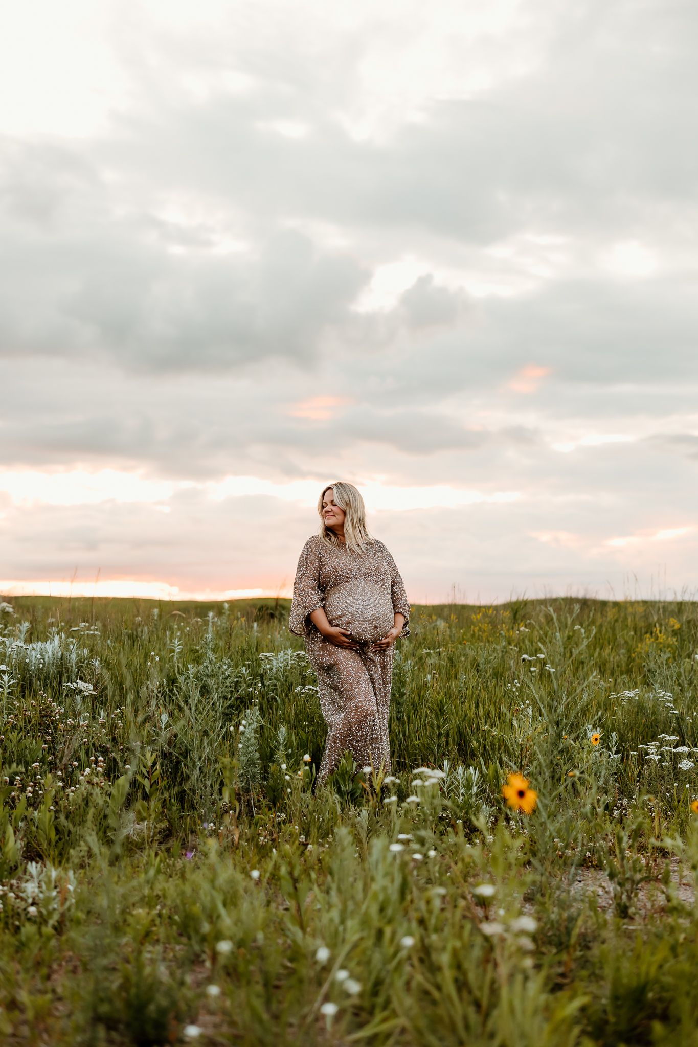 Pregnant woman in floral jumpsuit standing in wildflower field at sunset with dramatic cloudy sky.