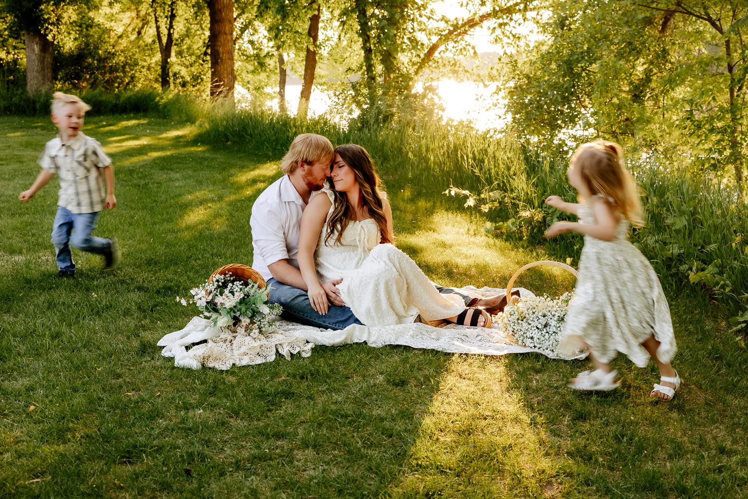 Family of four enjoying a sunny picnic on a blanket while children play around them outdoors.