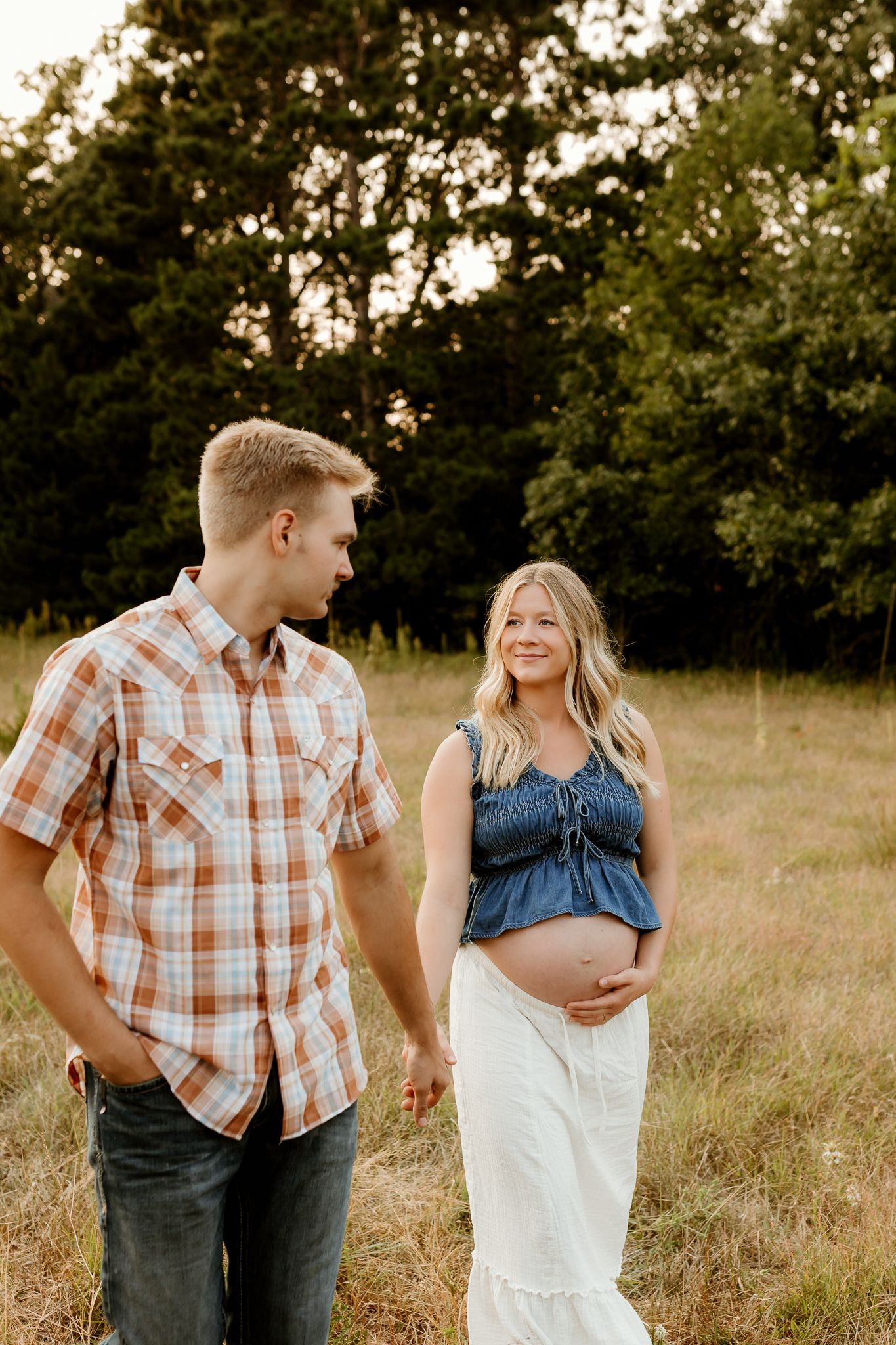 Pregnant woman in denim top and white skirt holds her baby bump while standing with her partner in a golden field.