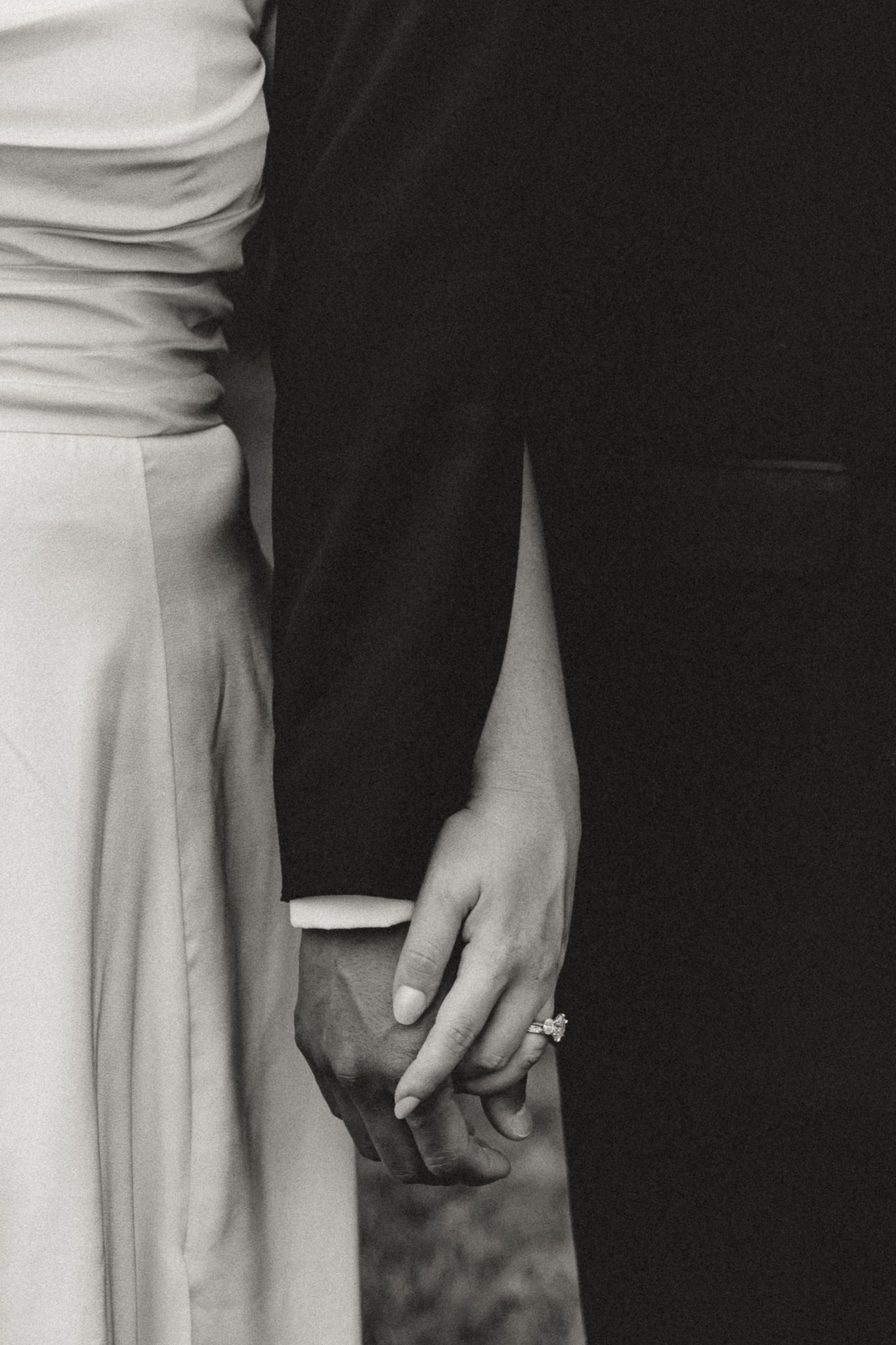 Black and white photo of a bride and groom holding hands, showcasing an engagement ring, in elegant wedding attire.