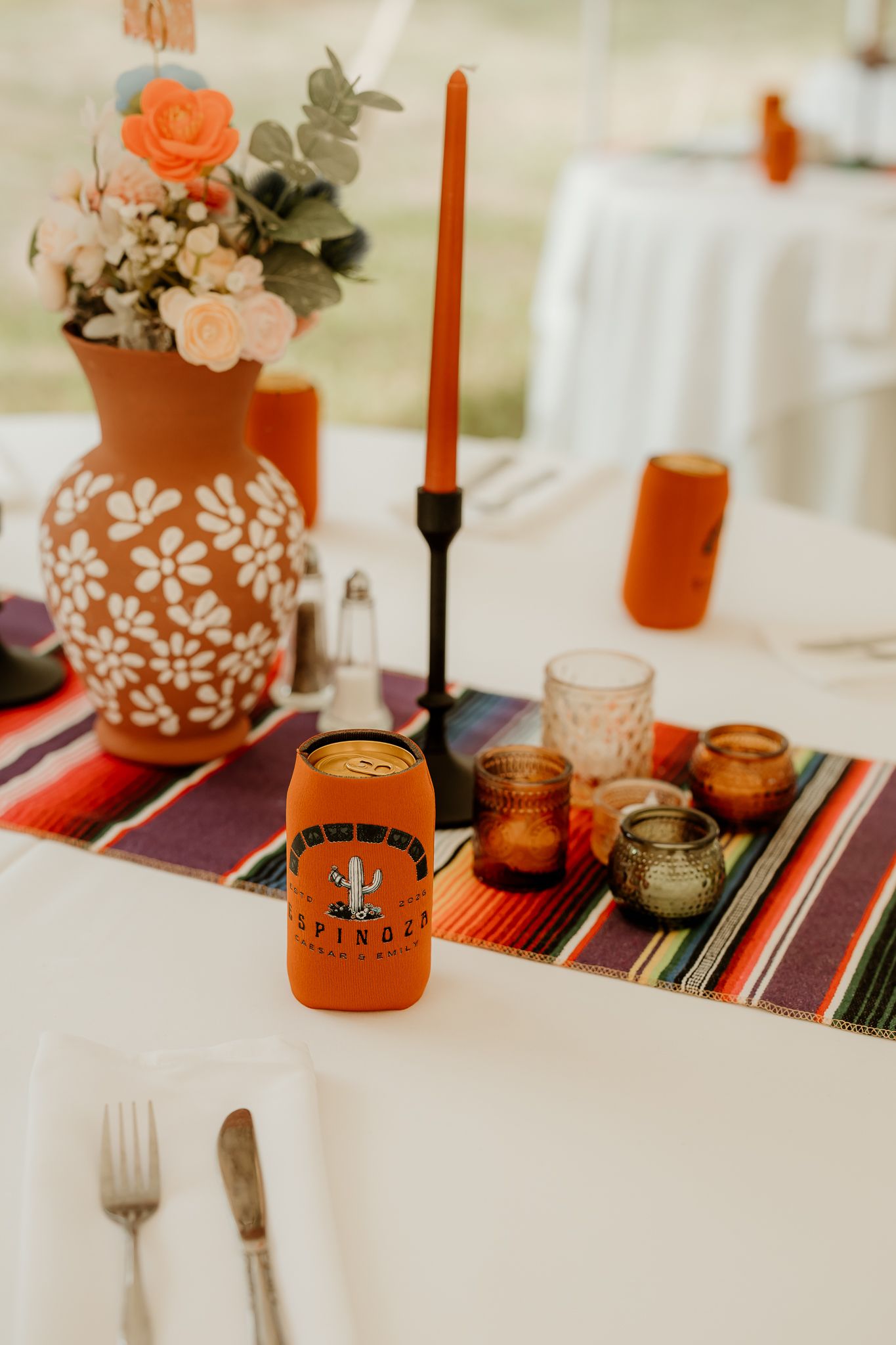 Colorful Mexican wedding table with orange floral vase, taper candle, votives, serape runner, and branded koozie centerpiece.