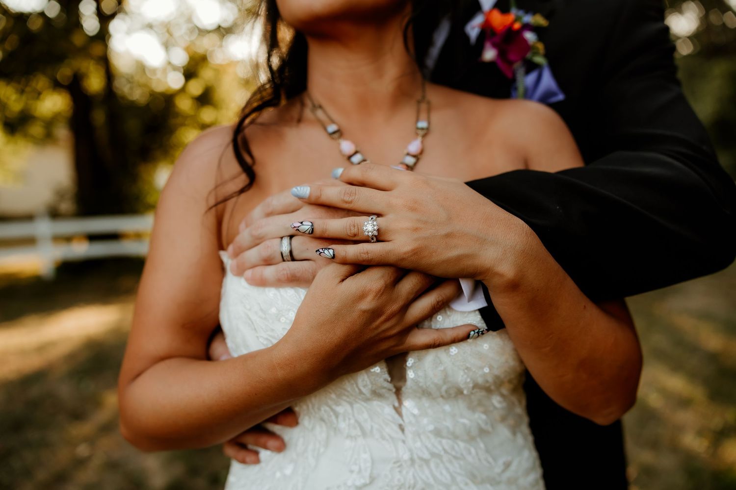 Bride in white lace dress with groom embracing her, showcasing wedding rings and turquoise necklace outdoors.