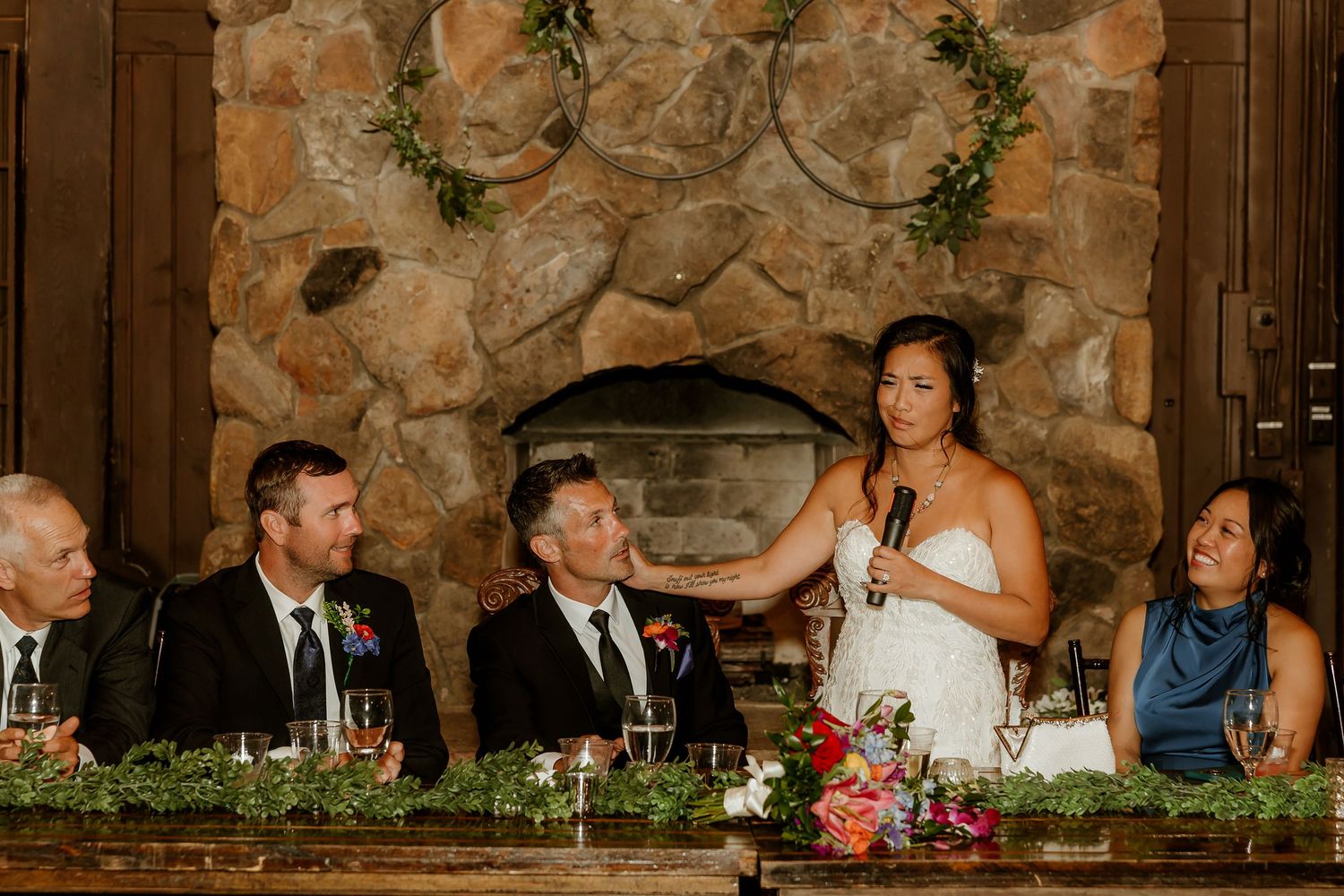 Bride gives a heartfelt speech at rustic stone fireplace wedding reception with guests seated at decorated head table.