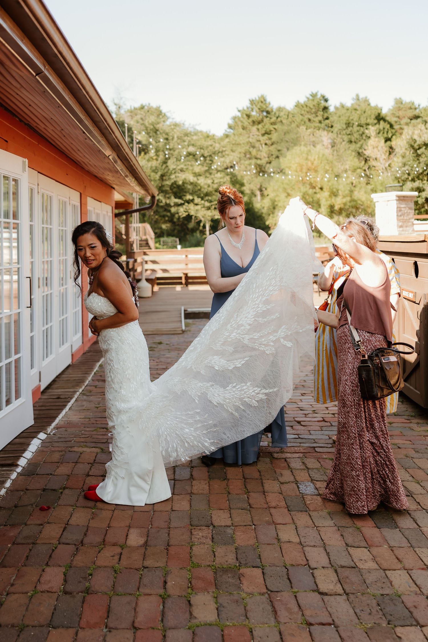 Bride in white gown gets veil adjusted by bridesmaids on brick patio outside rustic venue.