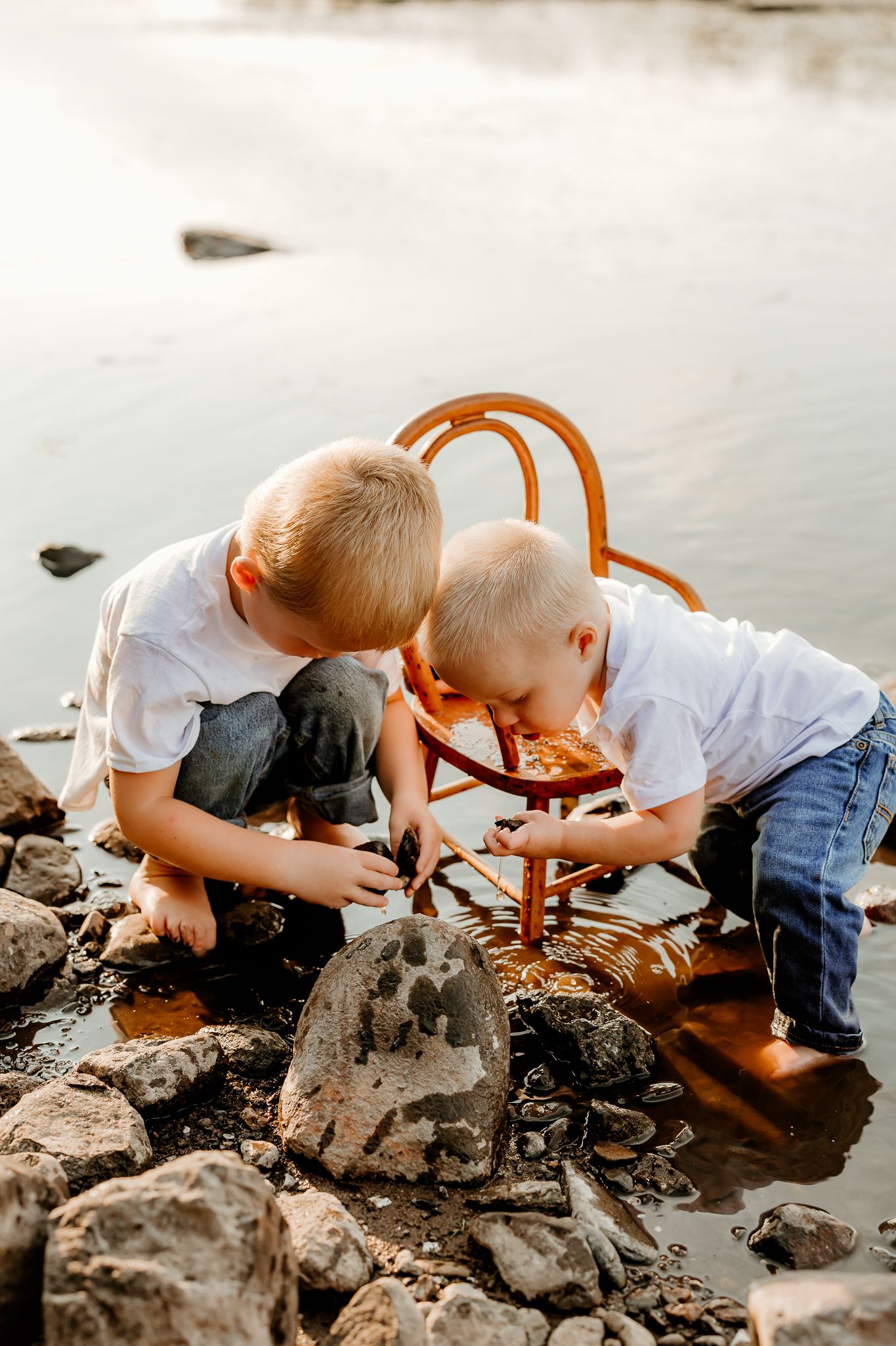 Two young boys in white shirts explore rocky shoreline together, crouching near water with a wooden chair nearby.