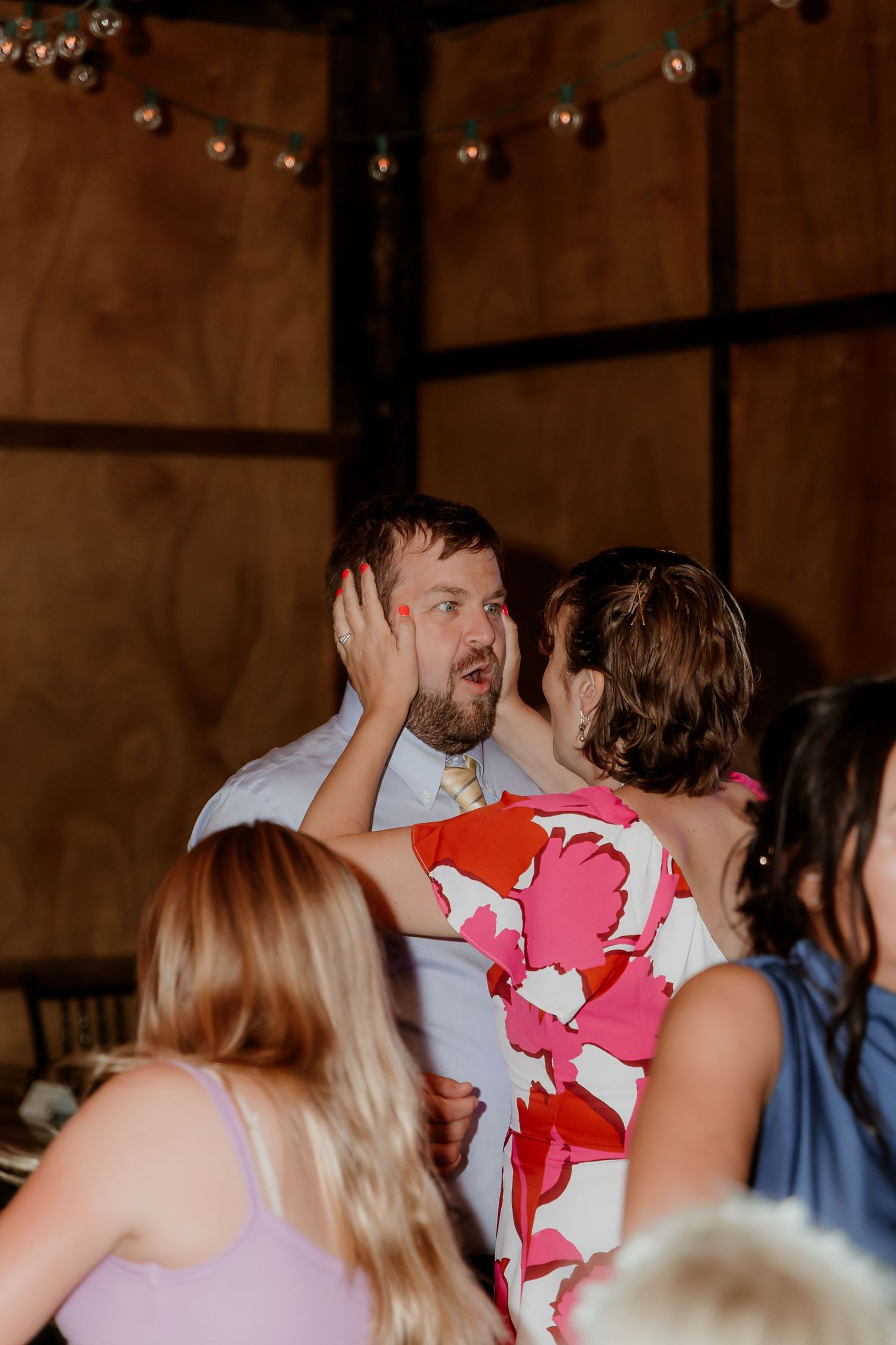 A man reacts with surprise as a woman in a floral dress touches his face at an indoor wedding reception.