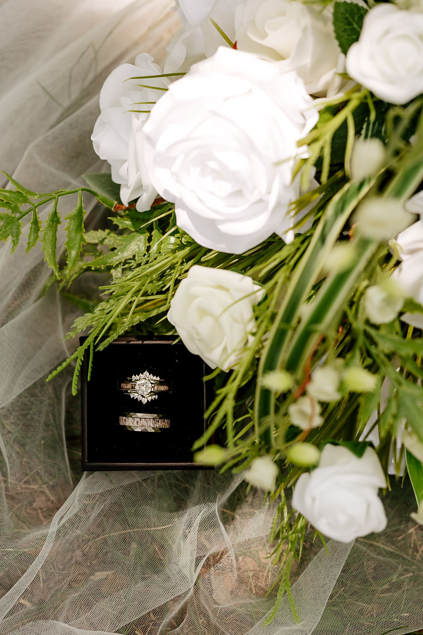 Wedding rings in black velvet box nestled among white roses and greenery on bridal bouquet with sheer veil.