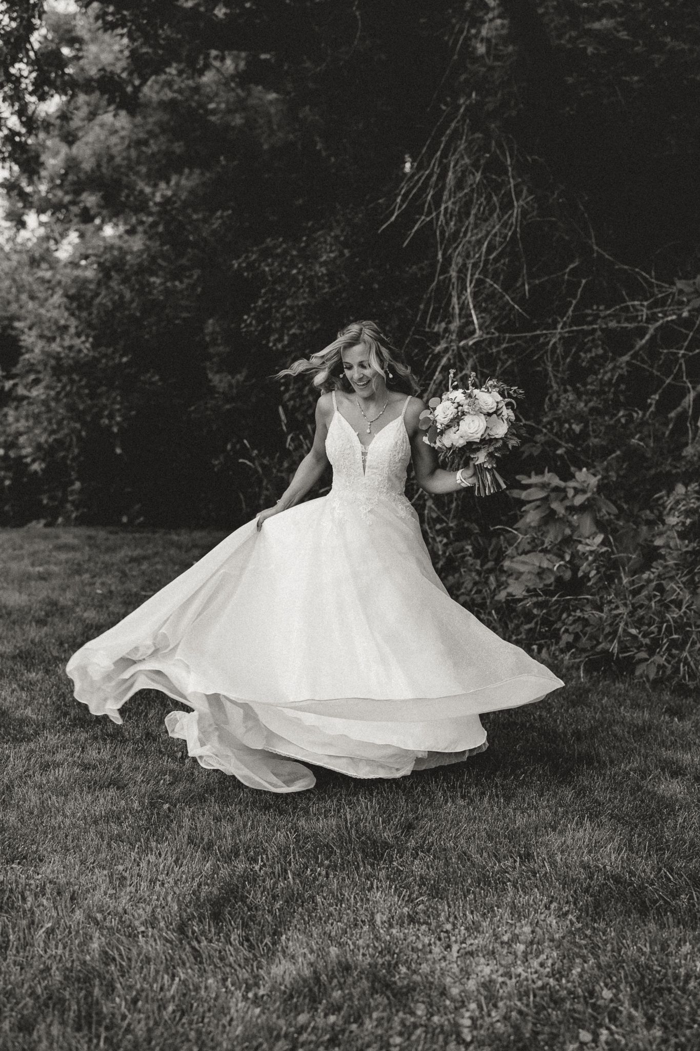 Bride twirling in a flowing white wedding gown outdoors, holding a bouquet, captured in elegant black and white photography.